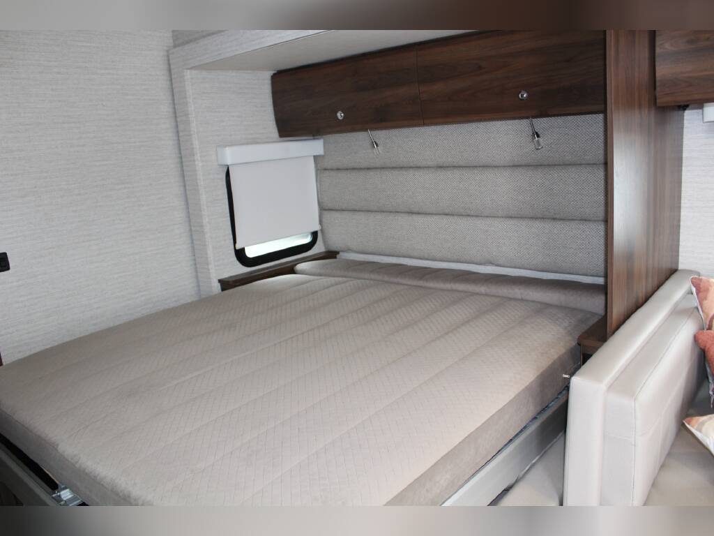 Cozy RV rental bedroom featuring a comfortable bed with a padded headboard, overhead storage, and a small window.. Winnebago Navion 2022