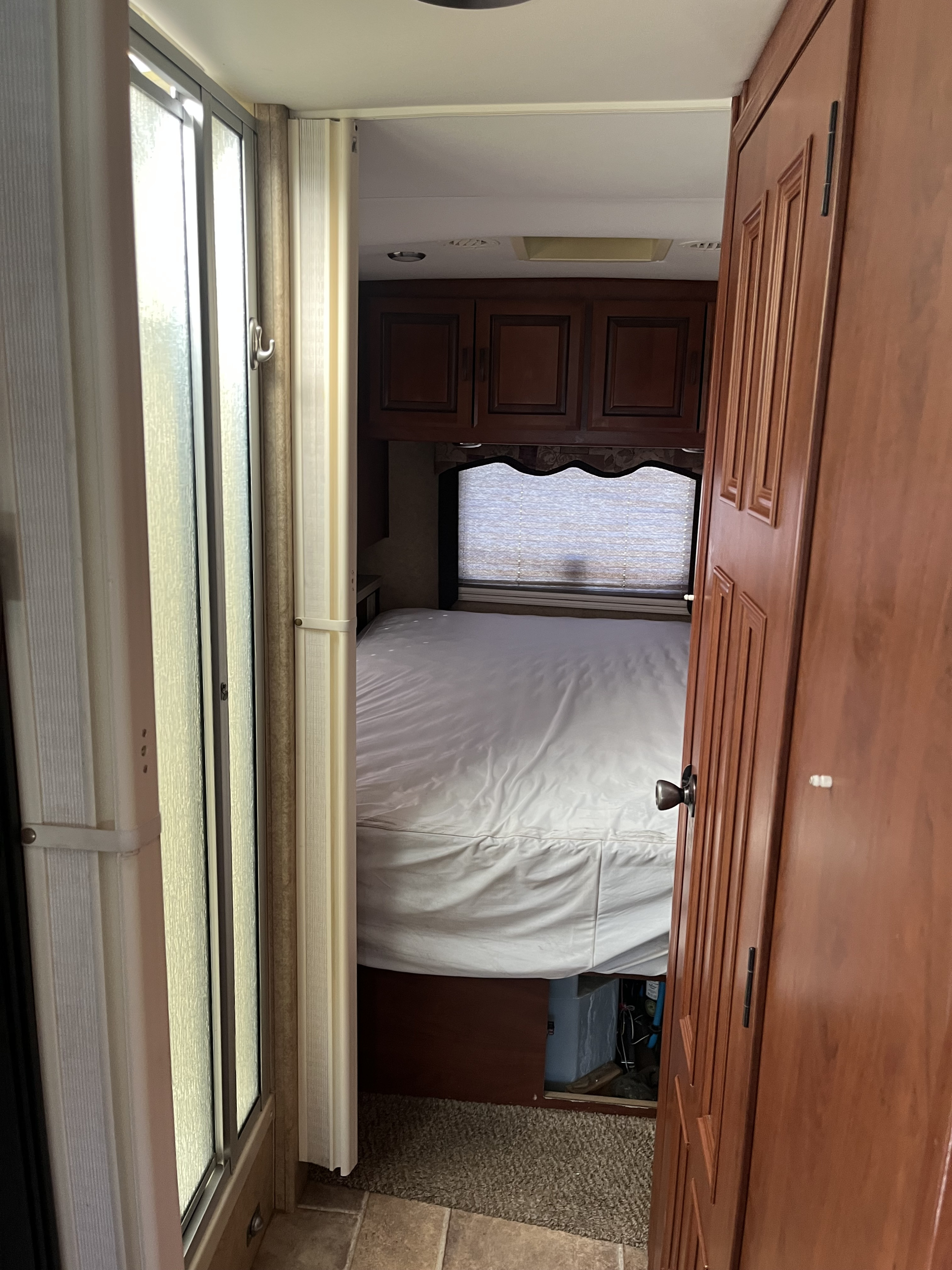 Inside an RV rental: a compact bedroom features a white bed, dark overhead cabinets, a window with blinds, and a shower on the left.. Lexington Grand Touring 2010