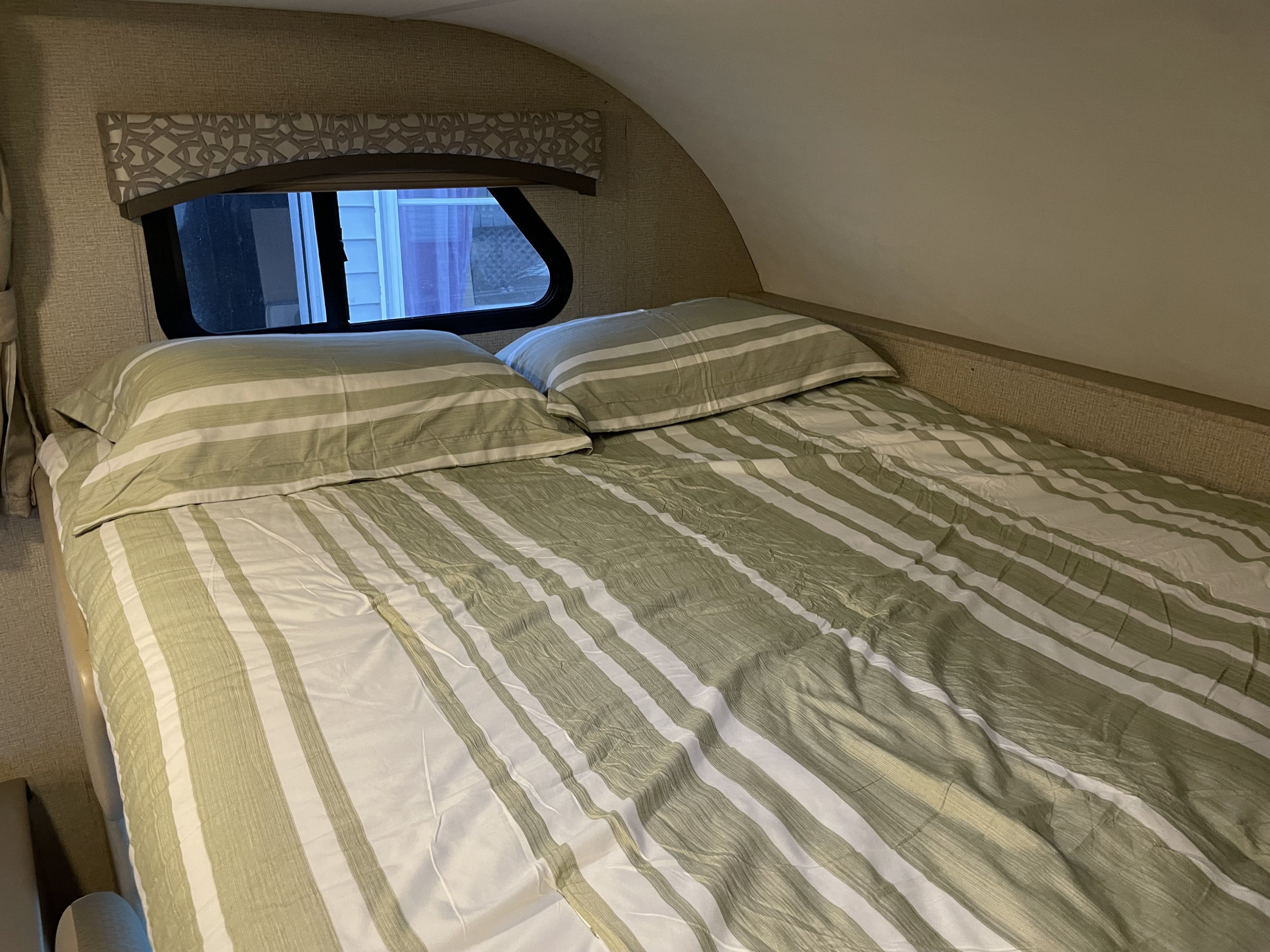 Inside an RV rental: a bed with green and white striped bedding, pillows, and a curved interior, visible through the window.. Thor Chateau 2018