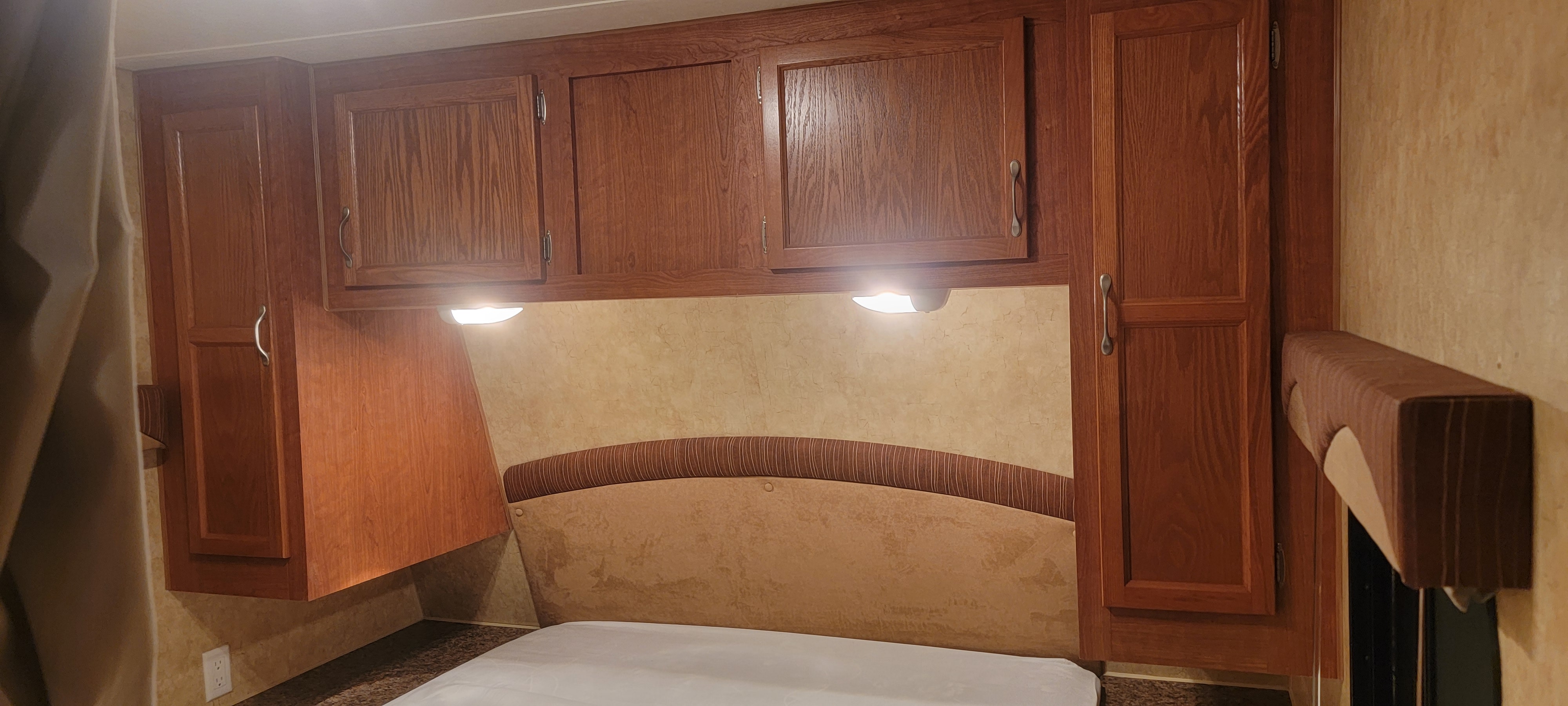 RV rental bedroom with overhead wood cabinets, upholstered headboard, and reading lights, offering a compact sleeping area.. Starcraft Autumn Ridge 2011