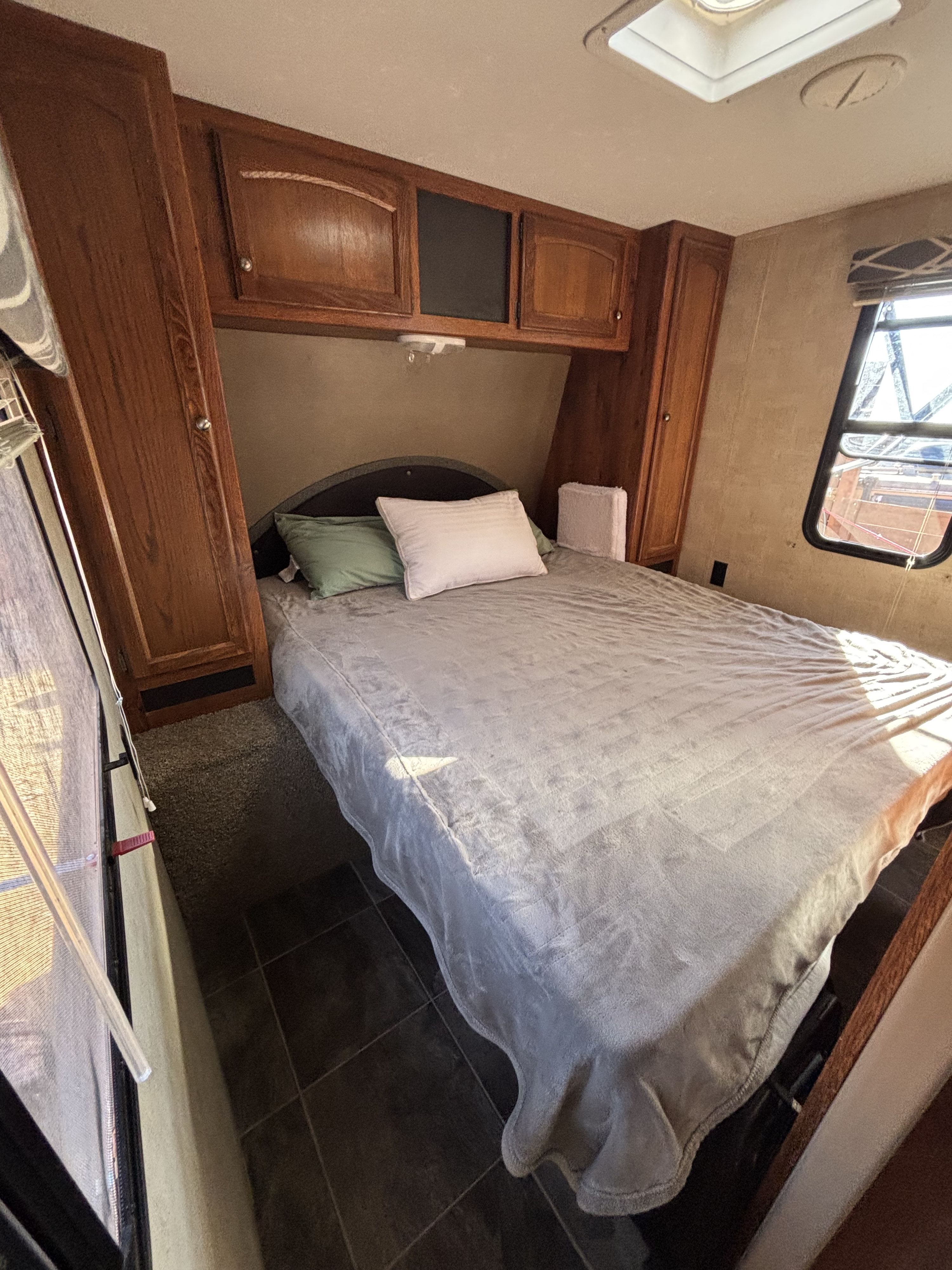 Spacious RV rental bedroom featuring a gray bed with pillows, wooden overhead cabinets, and a window for natural light.. Keystone RV Hideout Luxury 2015