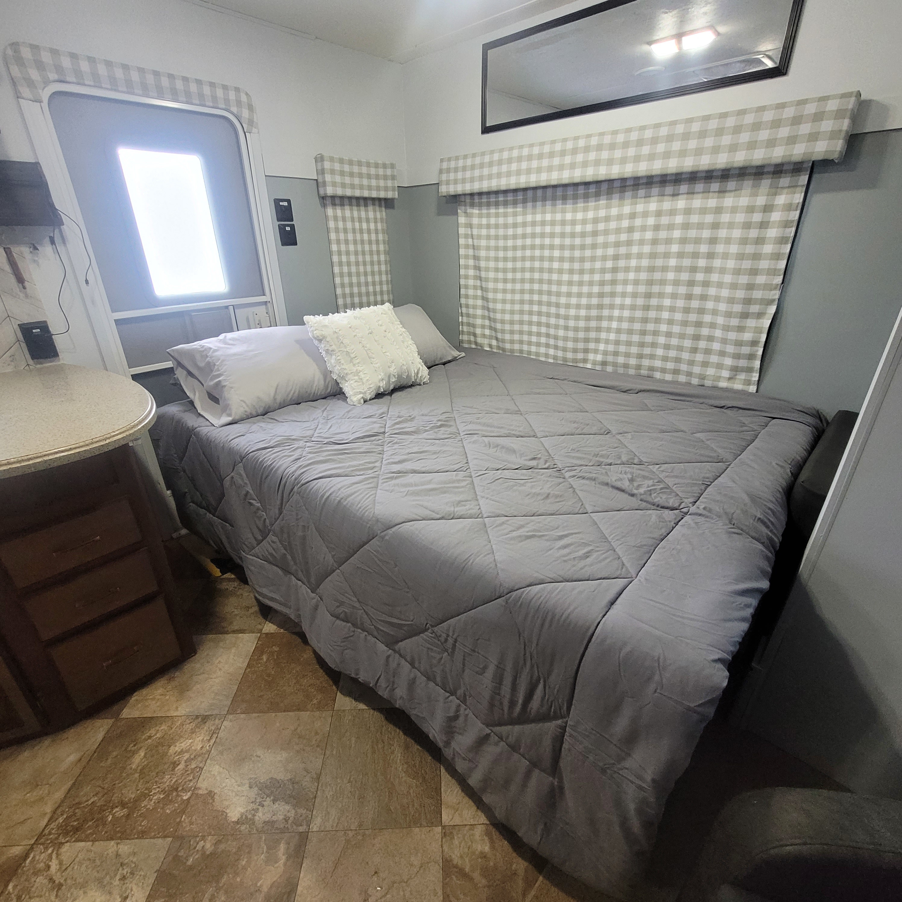 Compact RV rental bedroom featuring a comfy bed with grey bedding, checkered window treatments, and practical storage.. Crossroads RV Zinger 2014