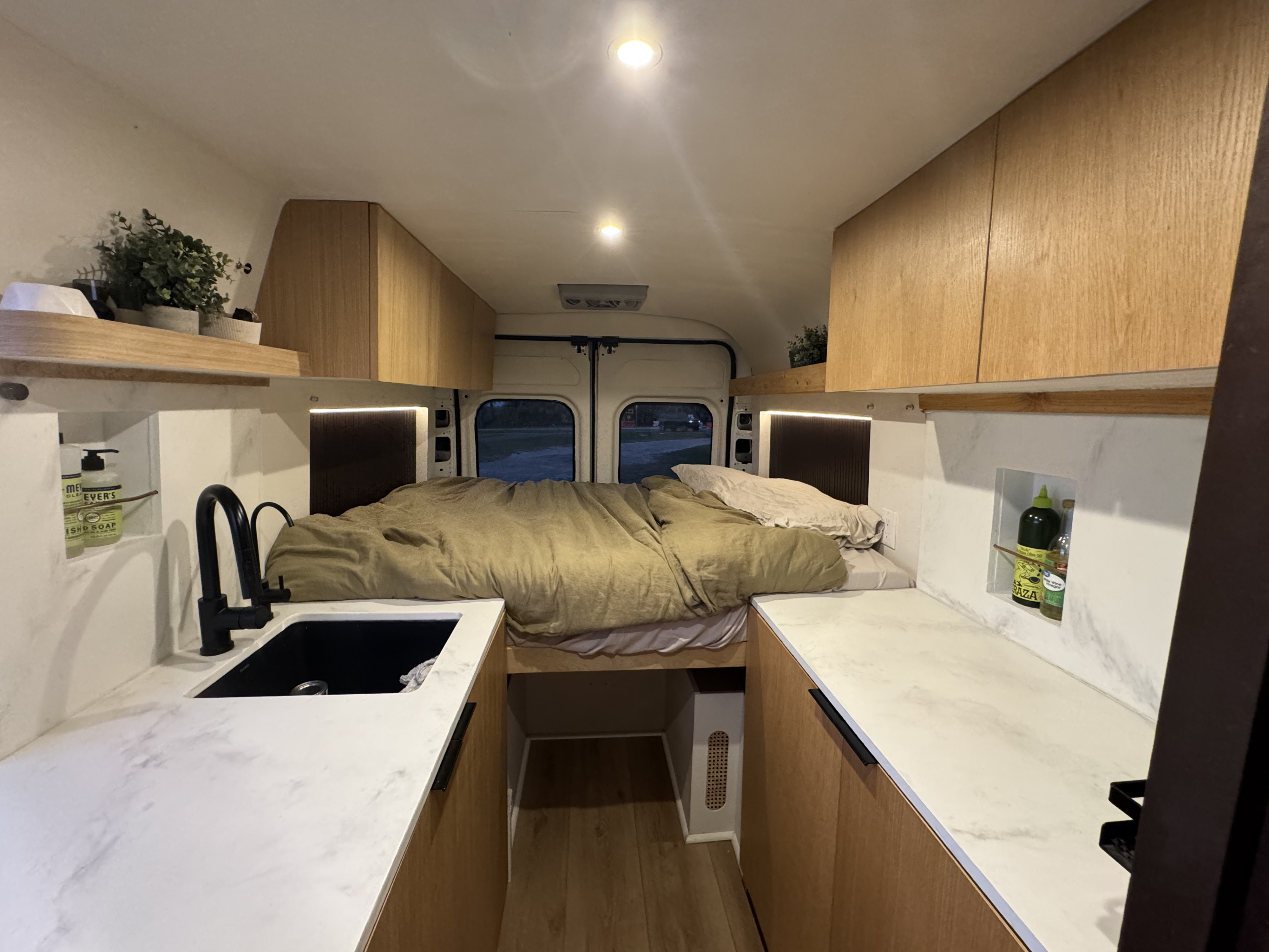 Interior of a stylish RV rental van conversion. Features a cozy bed, modern kitchen with white counters, and wood cabinets.. Dodge Promaster 2500 2021