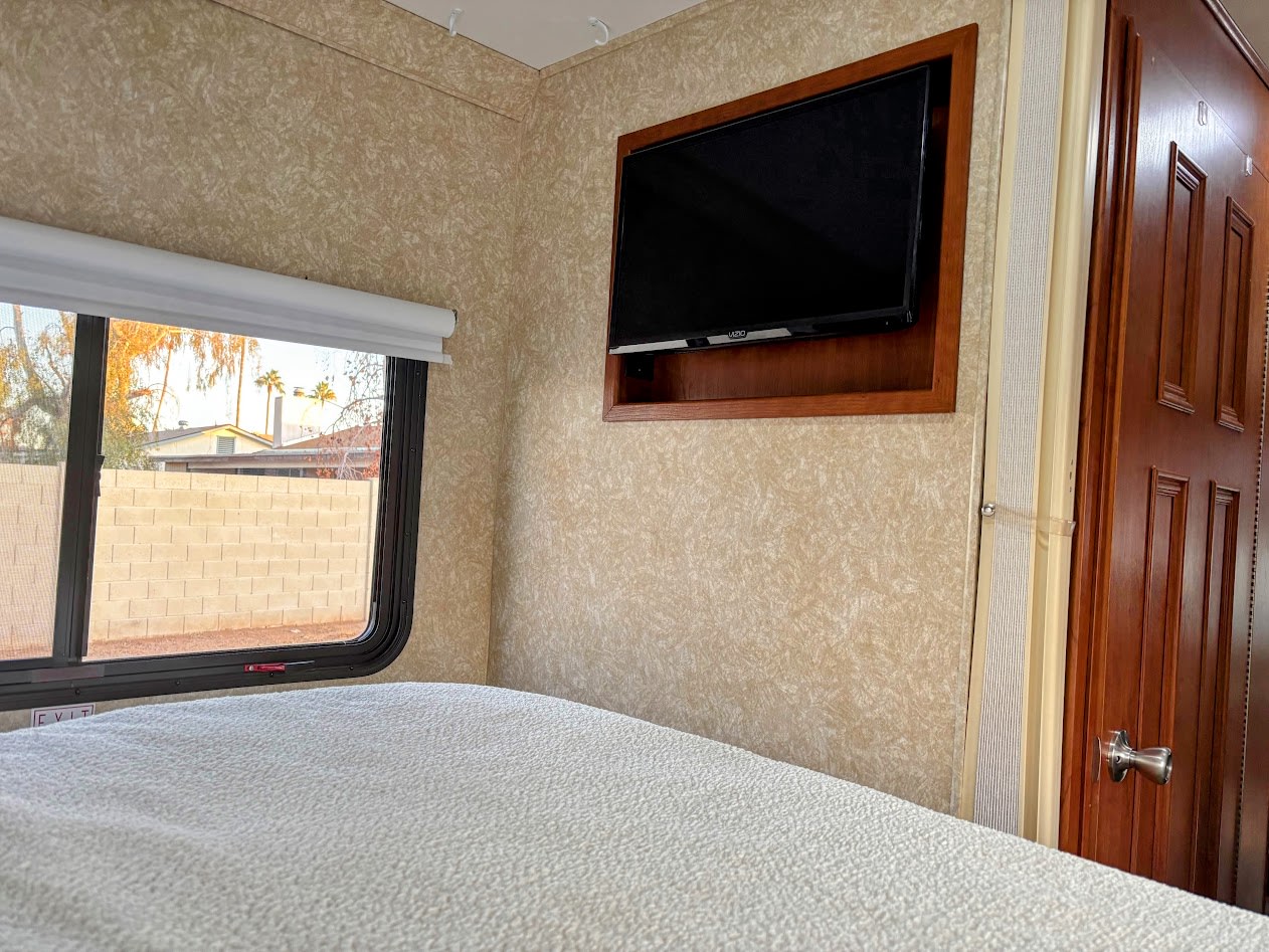RV rental bedroom with a white textured bed, large window showing outdoor view, and a TV mounted in wood on patterned walls.. Four Winds 5000 2008