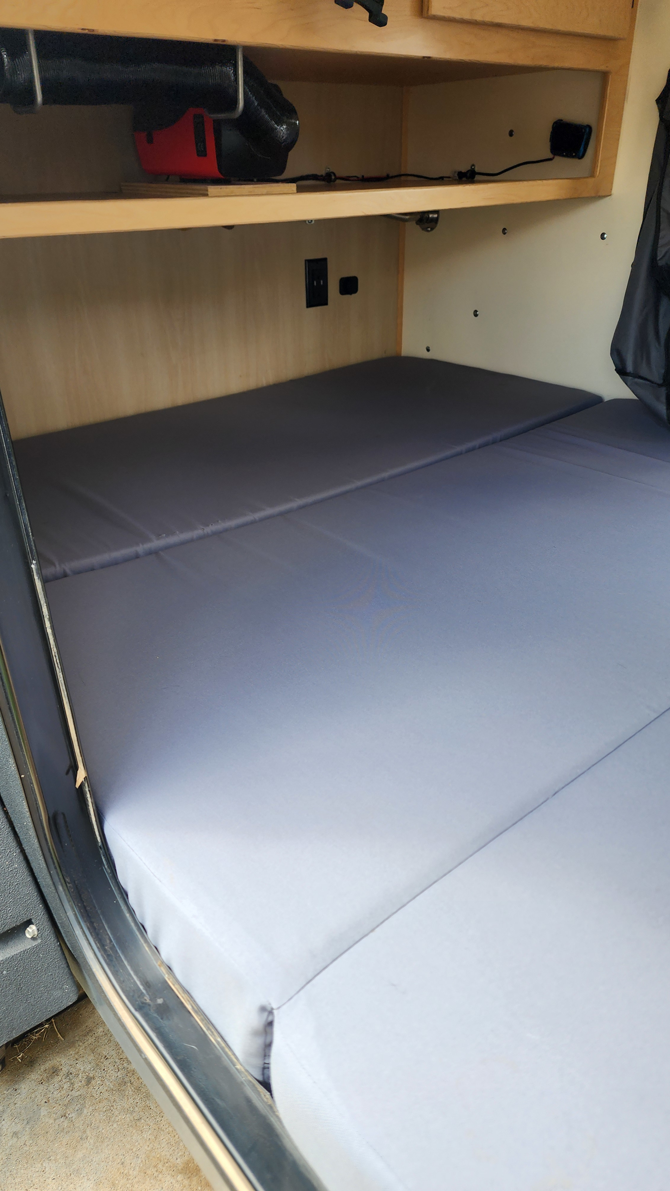 Inside an RV rental: A simple bed with gray mattresses, wooden shelves, and a heater/ventilation system.. High Altitude XT50 Treardrop Camper 2022