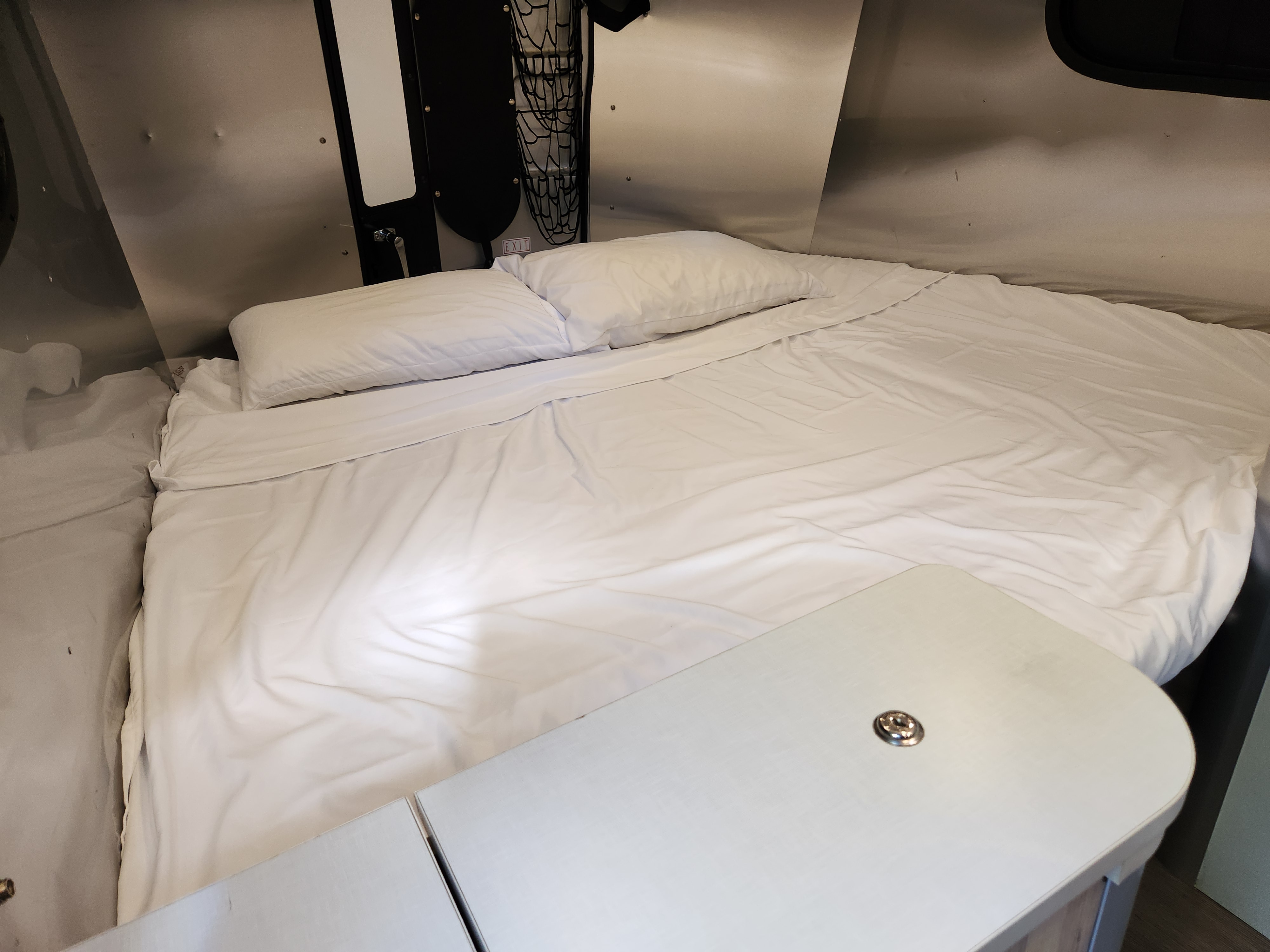 Cozy RV rental bed with white sheets & pillows. Compact interior featuring a table & wall storage.. Airstream Basecamp 2017