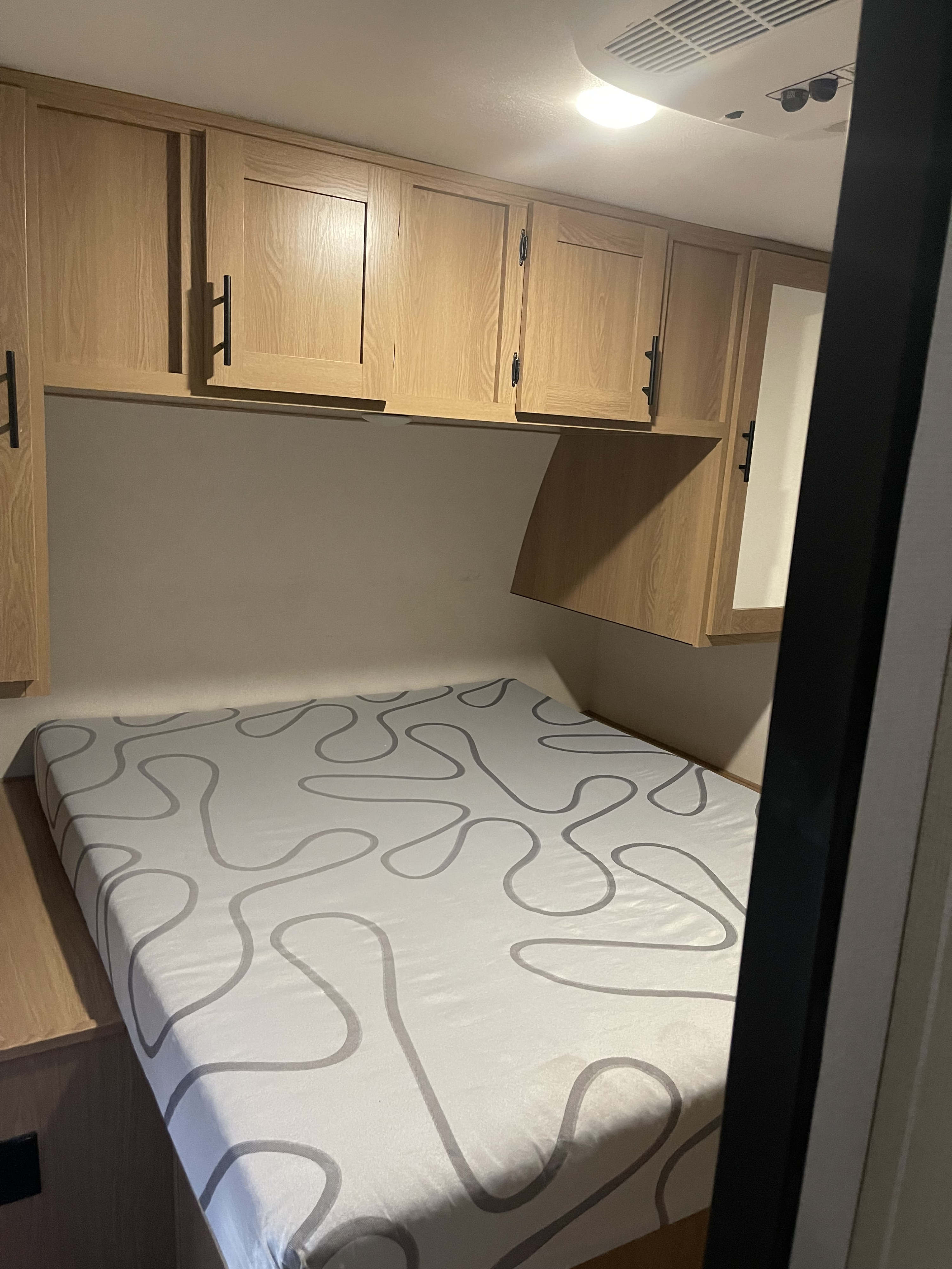 This RV rental interior: A compact bedroom with a bed featuring wavy grey patterns, light wood overhead cabinets, and a ceiling AC unit.. Avenger M28-BHS 2022