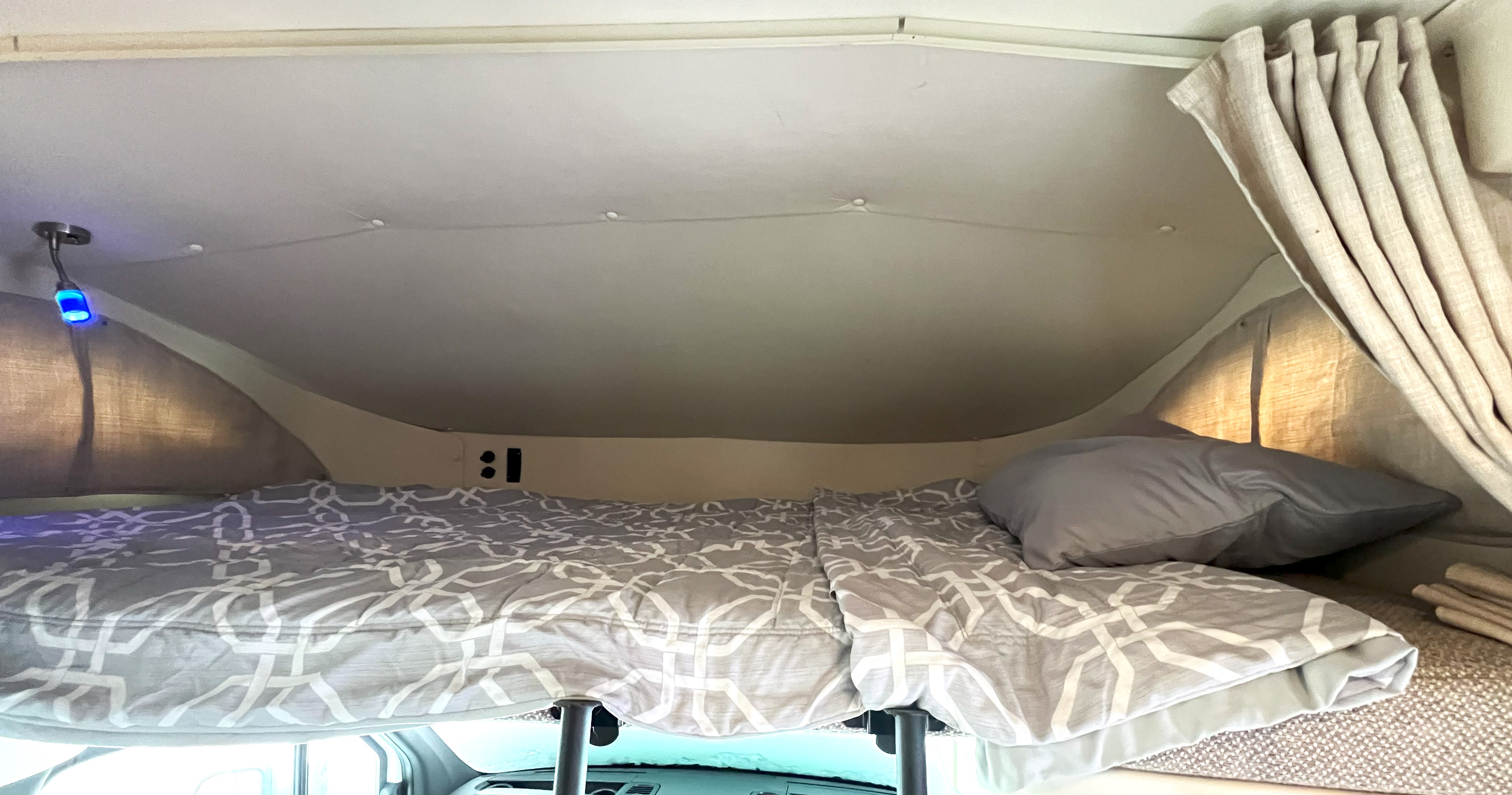 Cozy RV rental cab-over bed with patterned bedding, blue reading light, and curtains for comfortable travel.. Winnebago Navion 24D 2021
