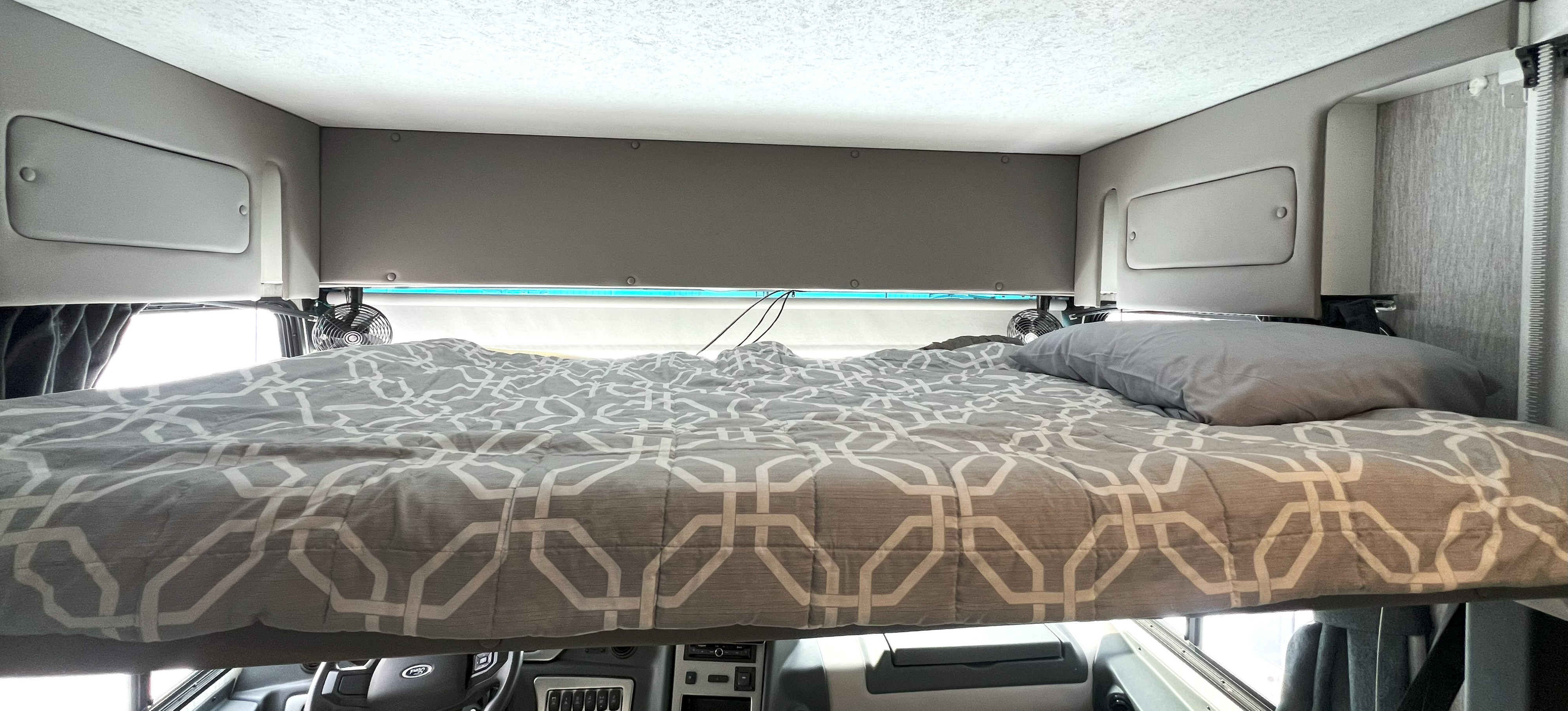 Overhead bunk bed in an RV rental, showing patterned bedding, a pillow, and cabin storage above the driver's area.. Coachmen Encore 2021