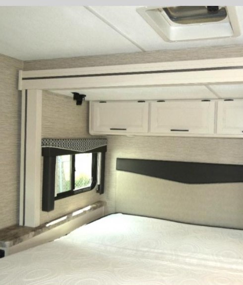 Inside this RV rental, find a compact bedroom with a comfortable bed, wall-mounted headboard, overhead cabinets, and window.. Thor Four Winds 2023