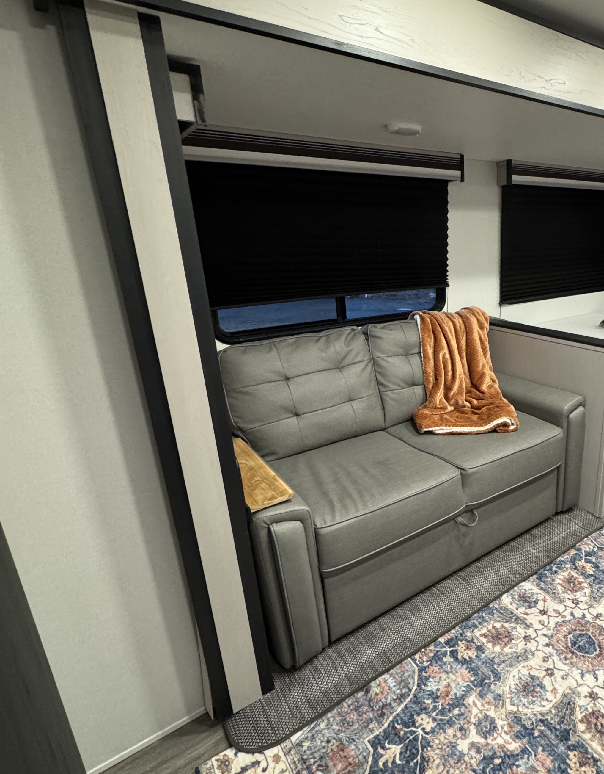 This RV rental features a cozy gray pull-out sofa with an orange blanket, black blinds, and a patterned rug on the floor.. Heartland RVs Mallard 2024