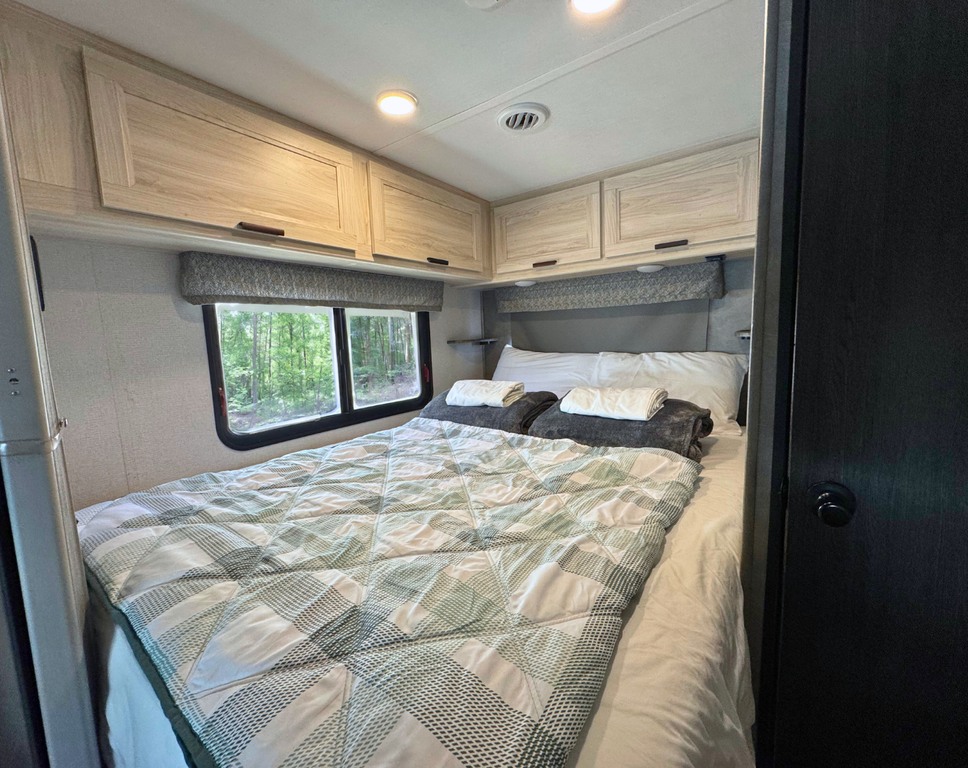 Inside an RV rental: a cozy bedroom with overhead cabinets, a comfortable bed, and a window showcasing a forest view.. Forest River Forester 2022