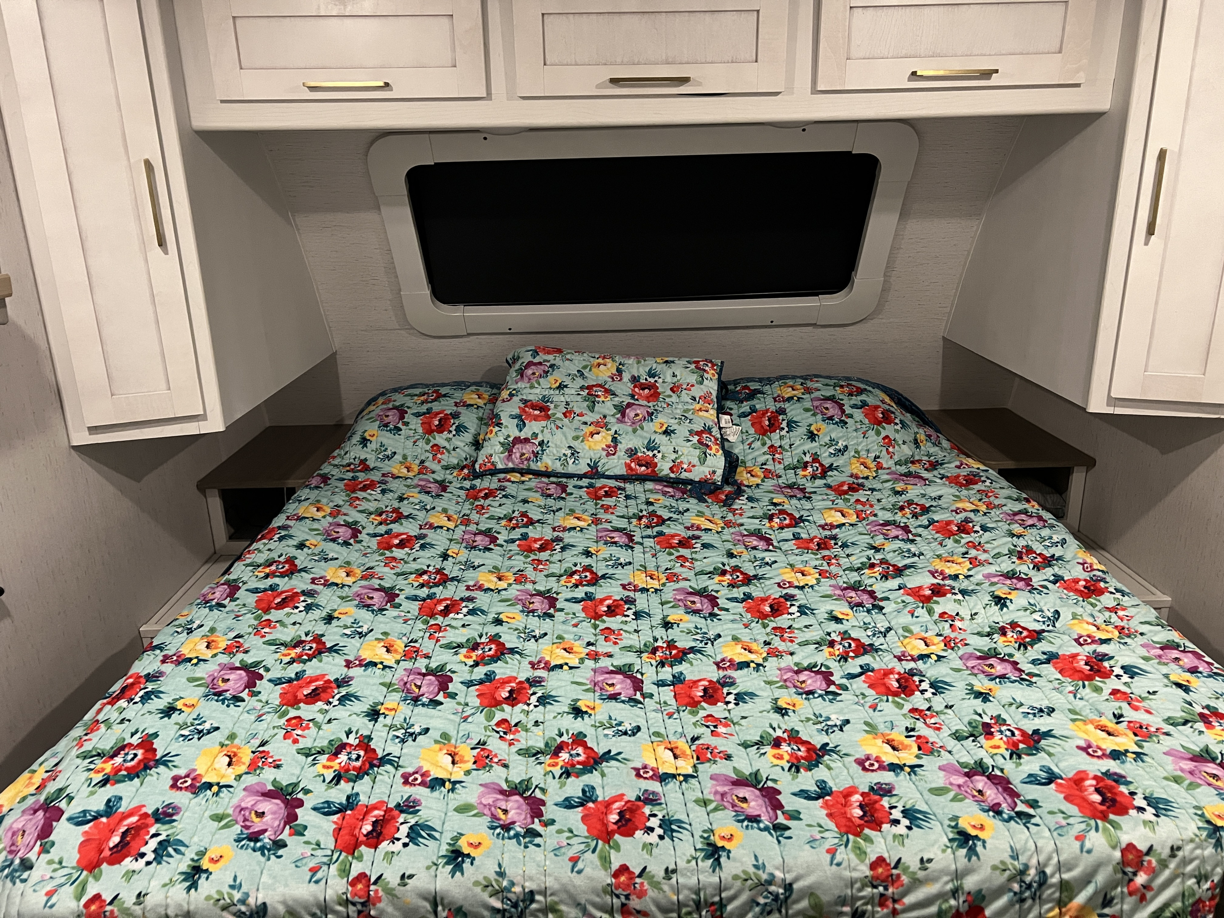 Cozy RV rental bedroom features a vibrant floral bed, overhead cabinets, and a unique window.. Astoria Astoria Trailer 2022