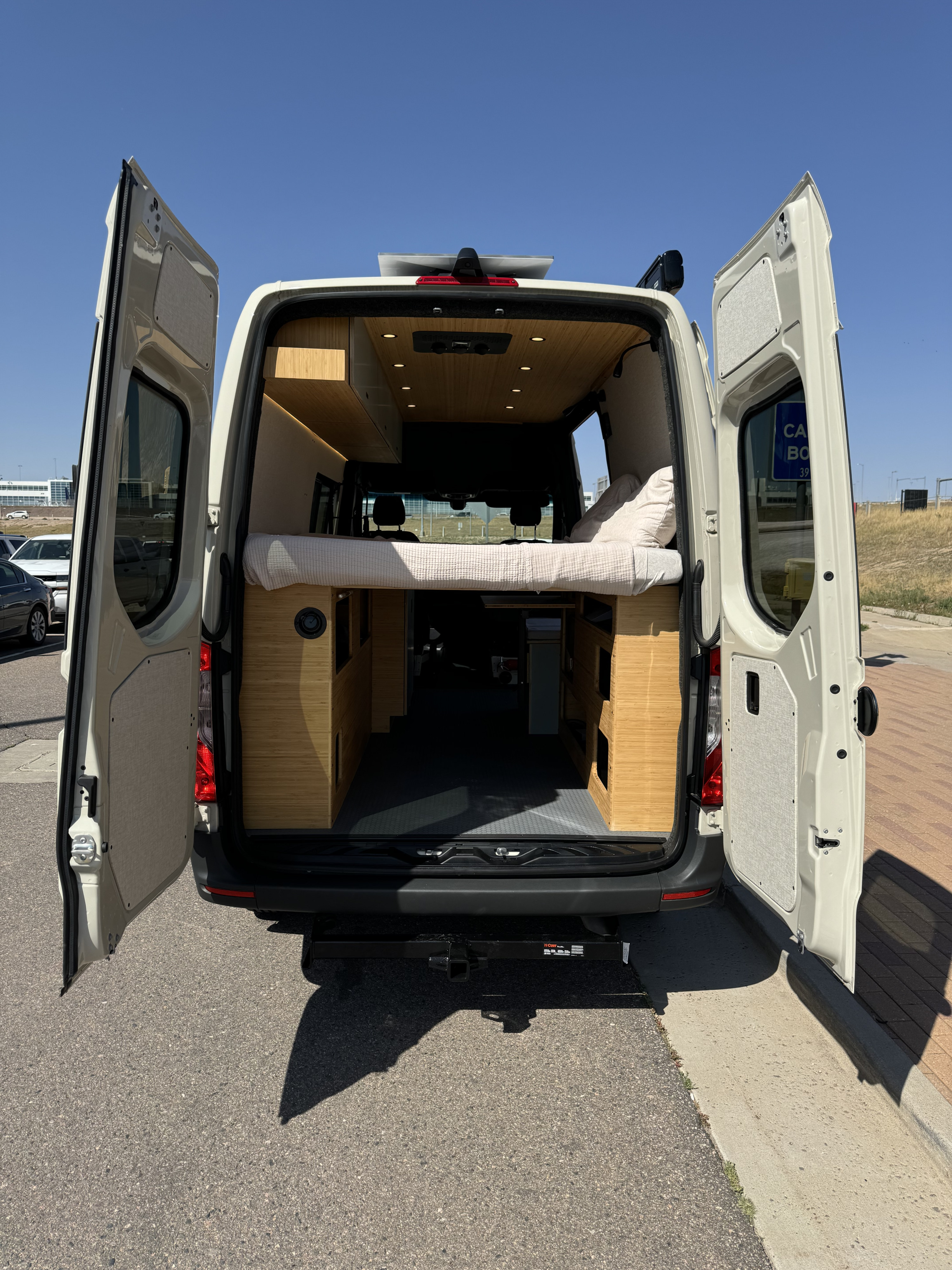Discover this RV rental camper van's custom interior: open doors reveal a cozy bed, wood cabinets, and ample lighting.. Mercedes-Benz Sprinter 2023