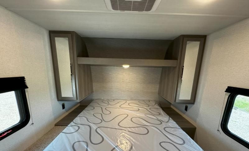 Inside an RV rental bedroom: a bed with patterned plastic covering, mirrored overhead cabinets, an upper shelf with a light, and side windows.. Keystone Hideout 2025