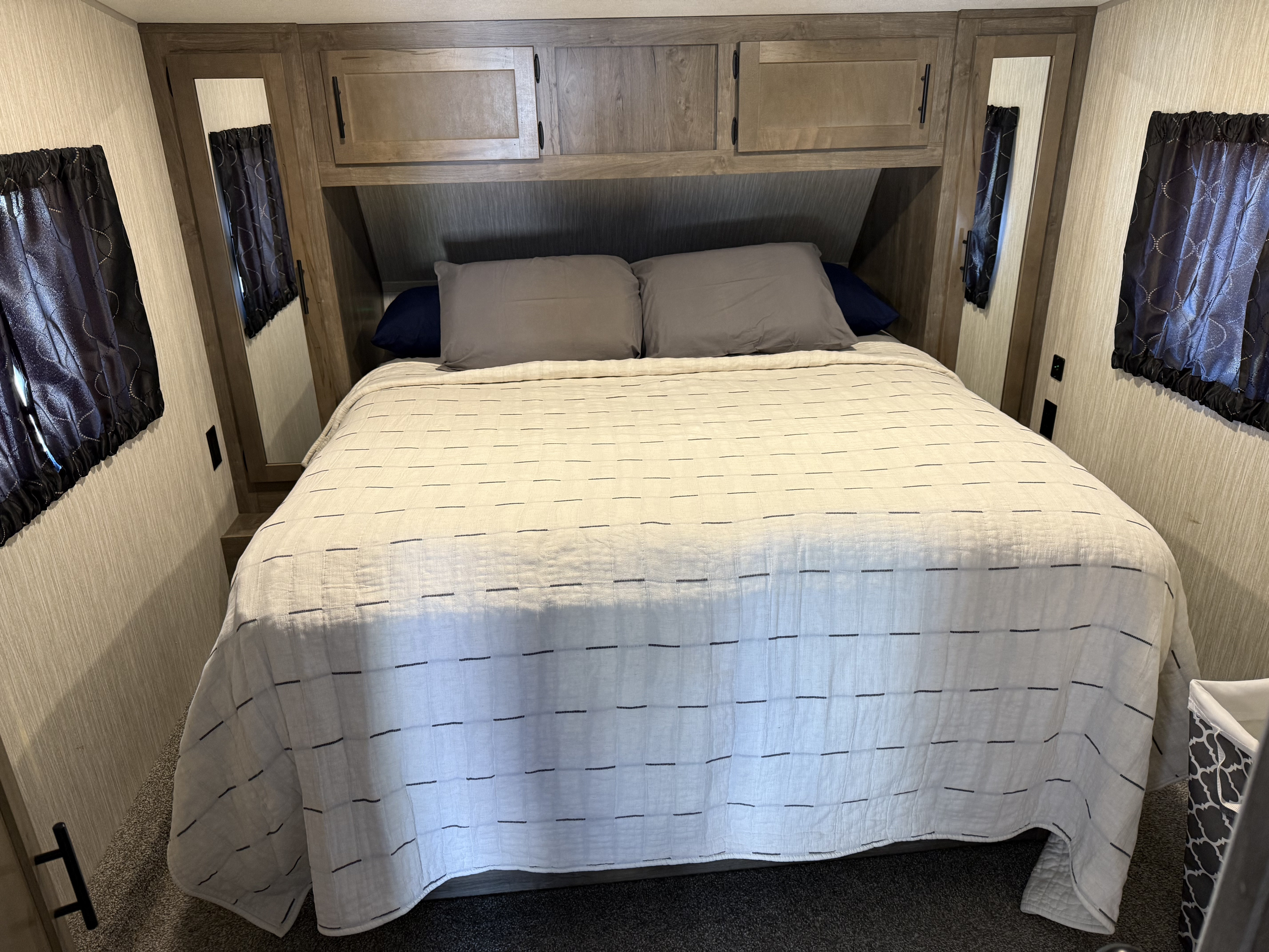 Cozy RV rental bedroom with queen bed, gray pillows, white quilt, wood cabinets, and dark curtains.. Forest River Cherokee Wolf Pack Toy Hauler 2021