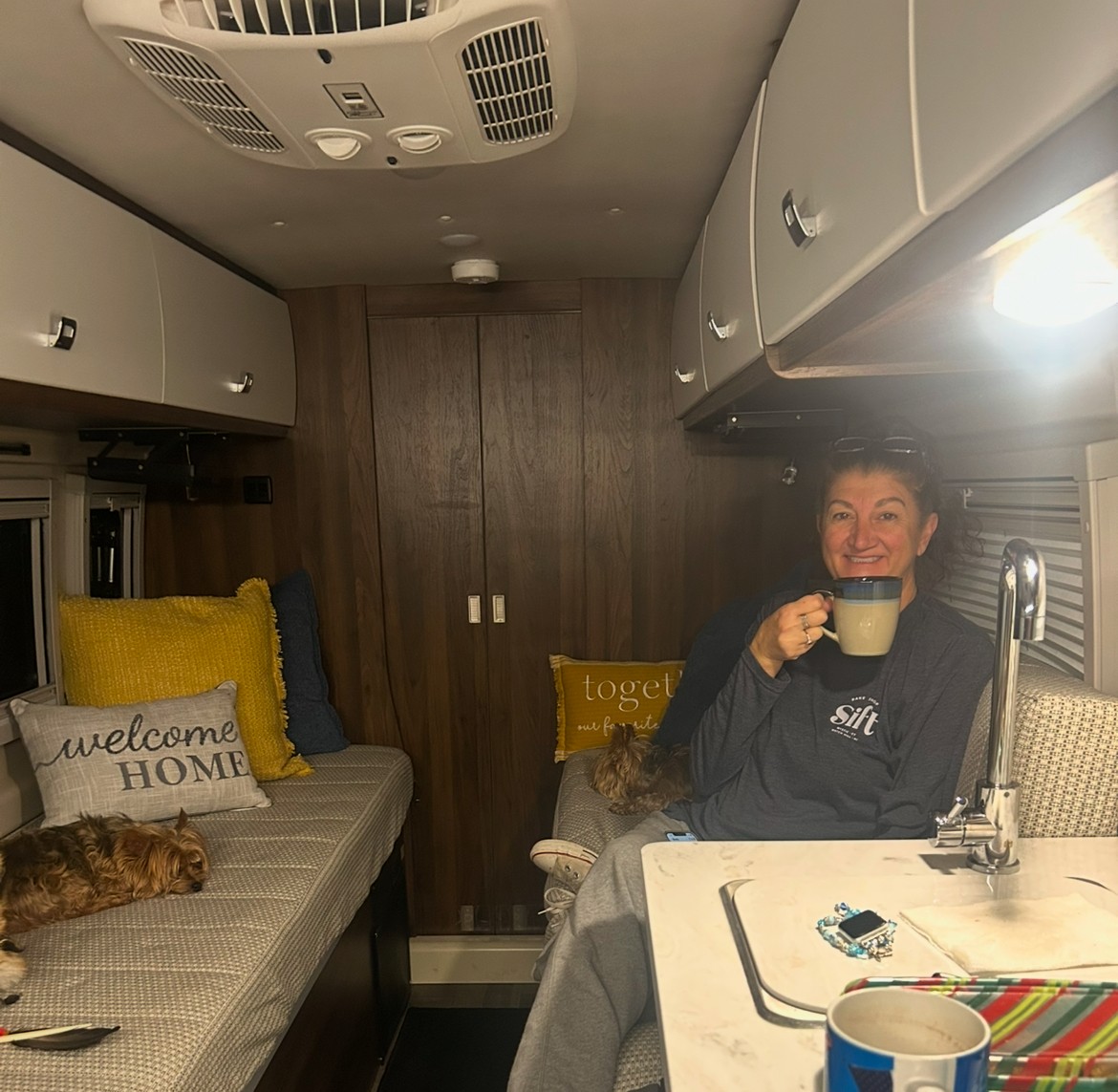 Smiling woman sips coffee with dogs in cozy RV rental. Ideal for travel getaways.. Winnebago Travato 2025
