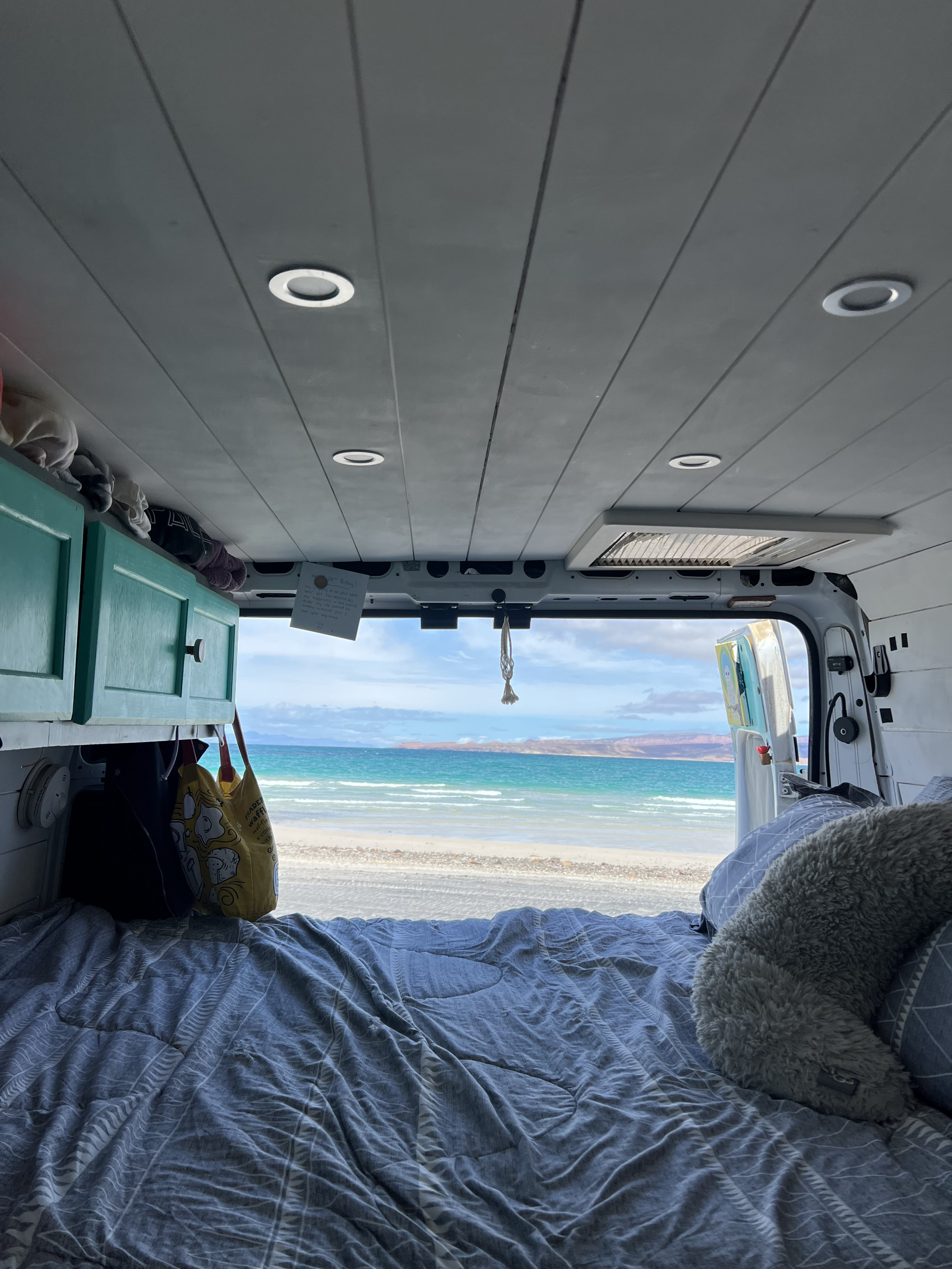 Cozy RV rental bed with a serene view of a sandy beach, turquoise ocean, and distant mountains. Enjoy the ultimate van life escape!. Ram Promaster 2500 2018