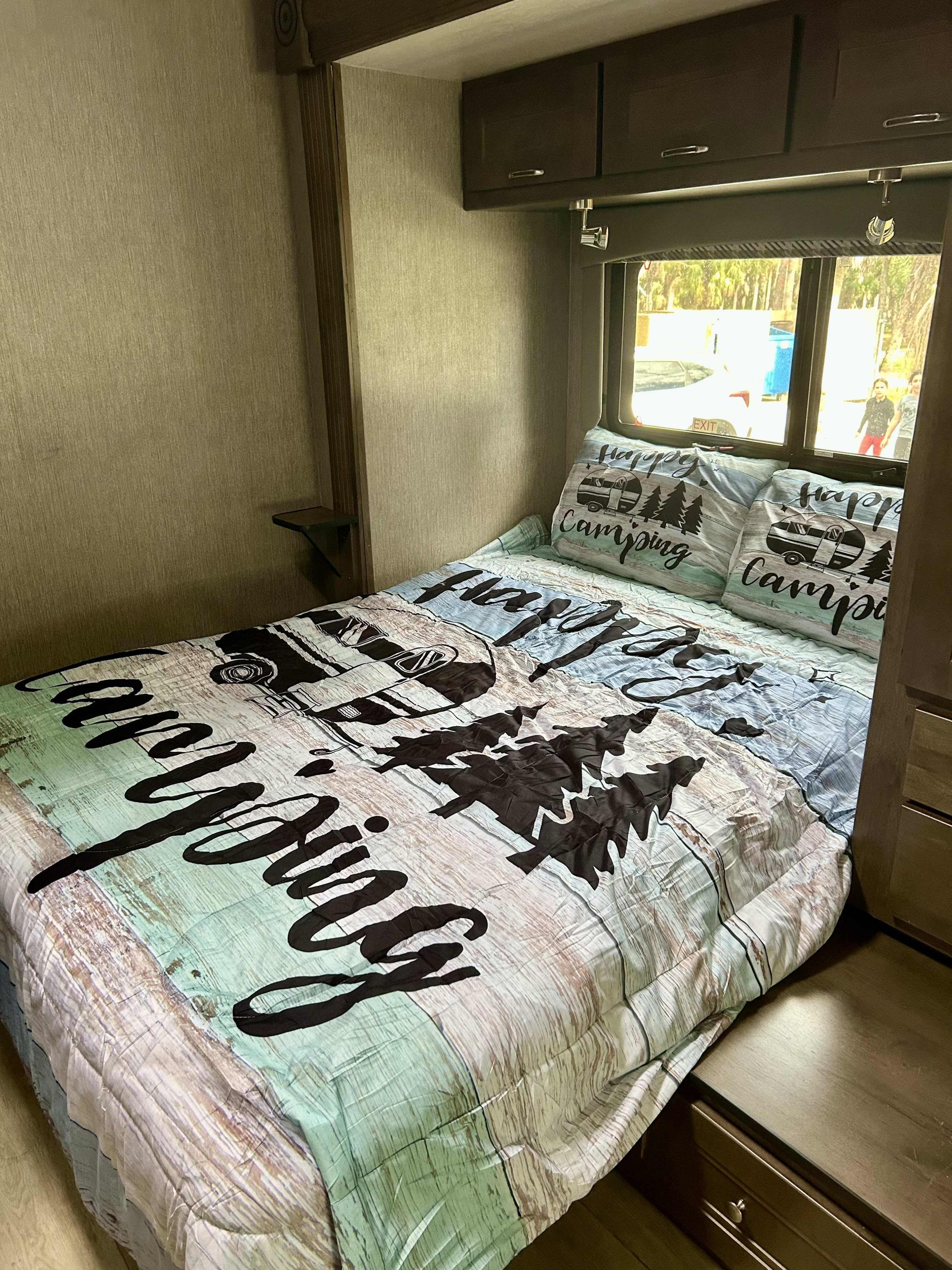 Cozy rv rental bedroom featuring a "Happy Camping" bed, overhead cabinets, and a window looking outside.. Nexus RV Phantom 2023