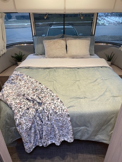 Cozy RV rental bedroom interior with a comfy bed, throw blanket, and plants. Curved windows show parked car.. Airstream Globetrotter 2021