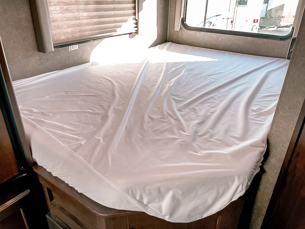 A neat RV rental bedroom features a bed with clean white sheets. Windows with blinds and an EXIT sign are visible.. Thor Motor Coach Freedome Elite 2020