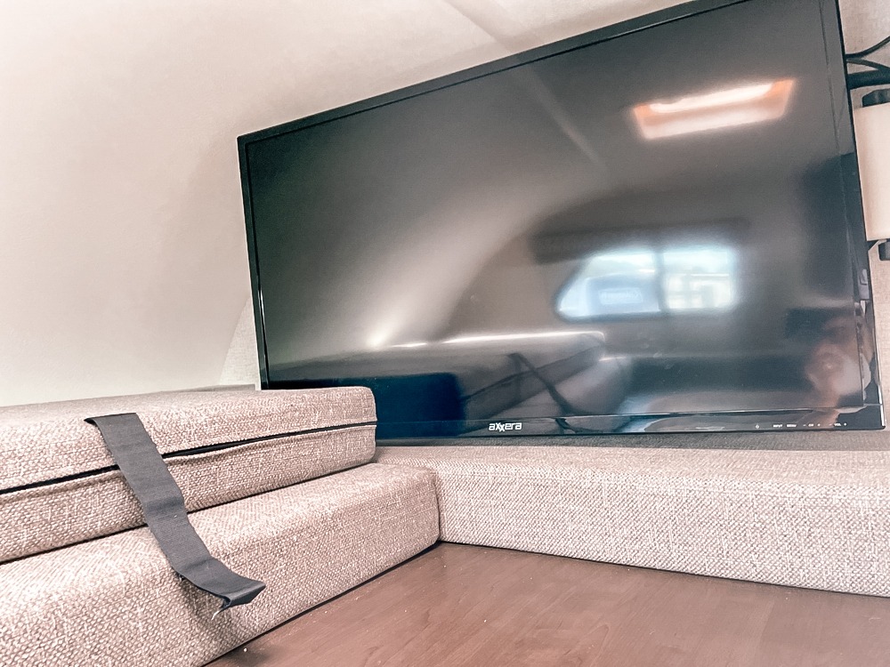 An RV rental's cozy corner features a flat-screen TV reflecting a window, next to stacked cushions with a strap.. Thor Motor Coach Freedome Elite 2020