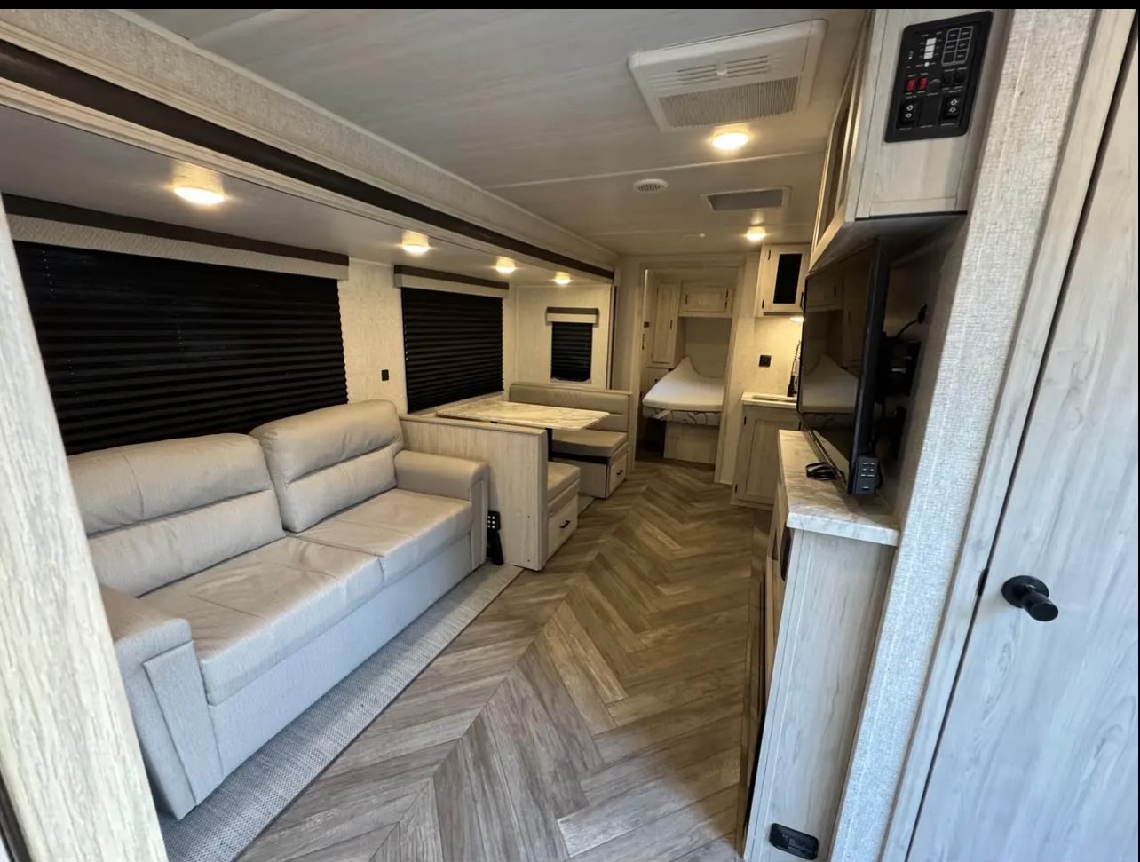 Modern RV rental interior: light sofa, dinette, kitchen with TV, and rear bedroom. Features light wood trim and herringbone floor.. East to West Della Terra 2022