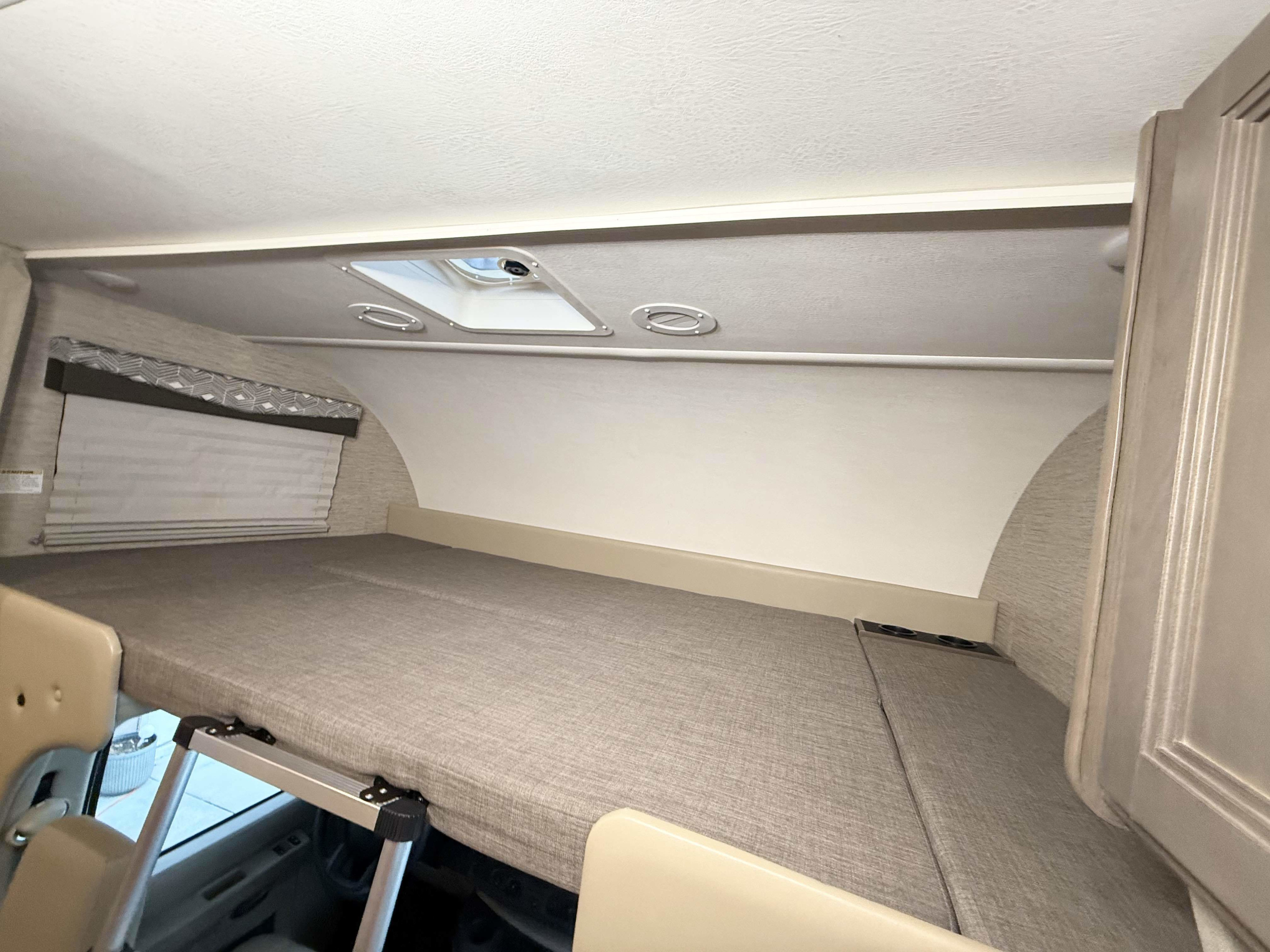 Inside an RV rental: the large over-cab bunk with a mattress, window shade, ceiling vent, and AC vents.. Thor Four Winds 2023