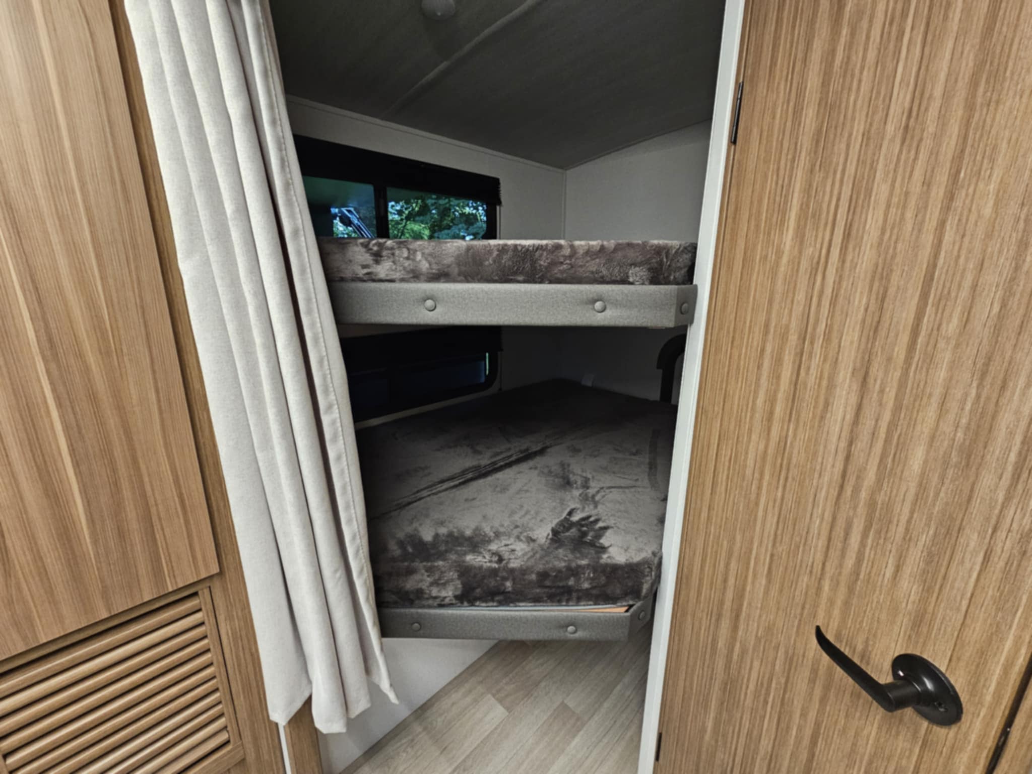 Inside an RV rental, showing the cozy bunk beds with soft grey blankets, light wood walls, and a small window.. Keystone RV Passport SL 2025