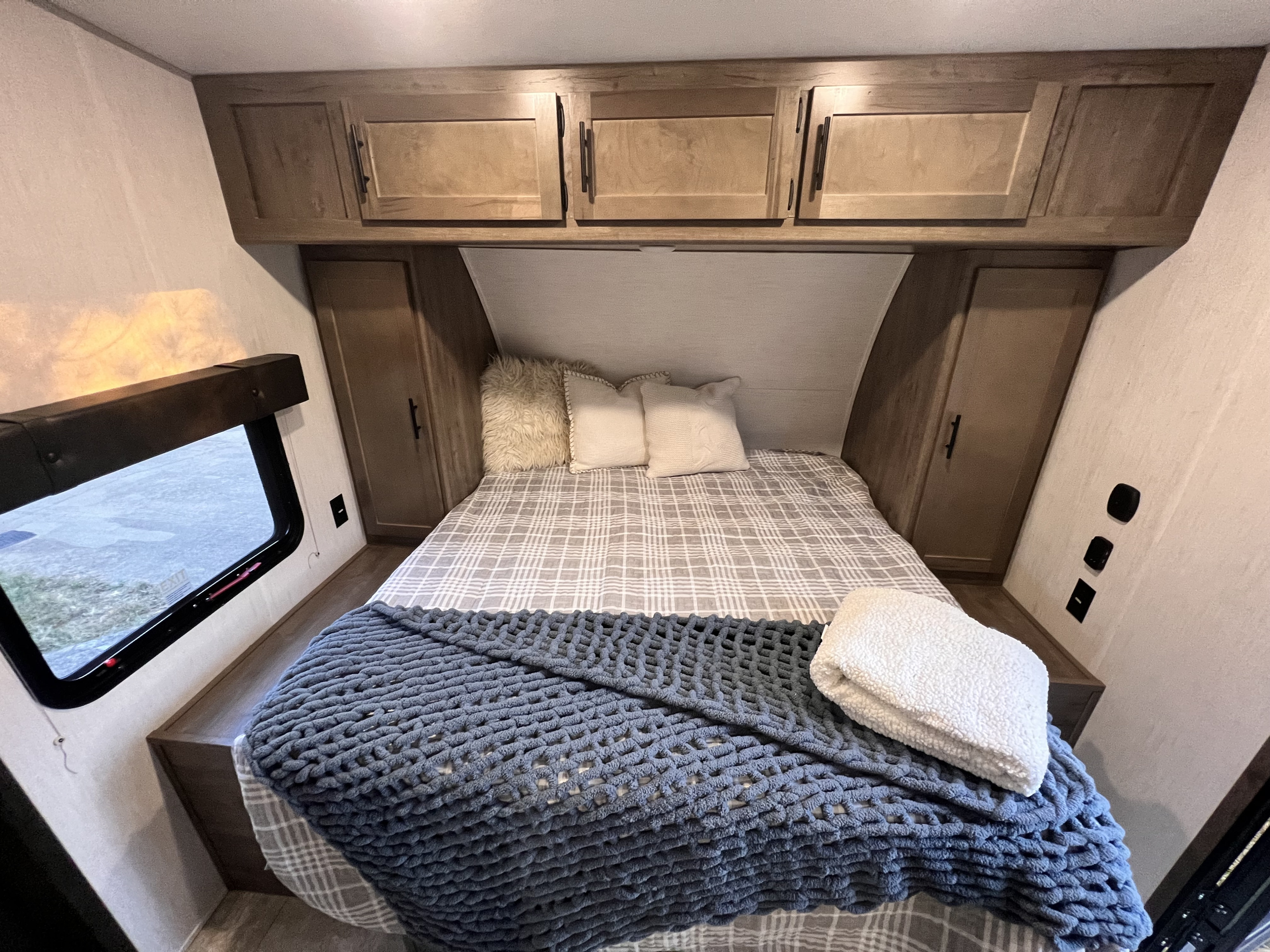 Cozy RV rental bedroom with a neatly made bed, featuring plaid bedding, a blue knit blanket, and overhead wood cabinets.. Forest River 29ks rouge 2022