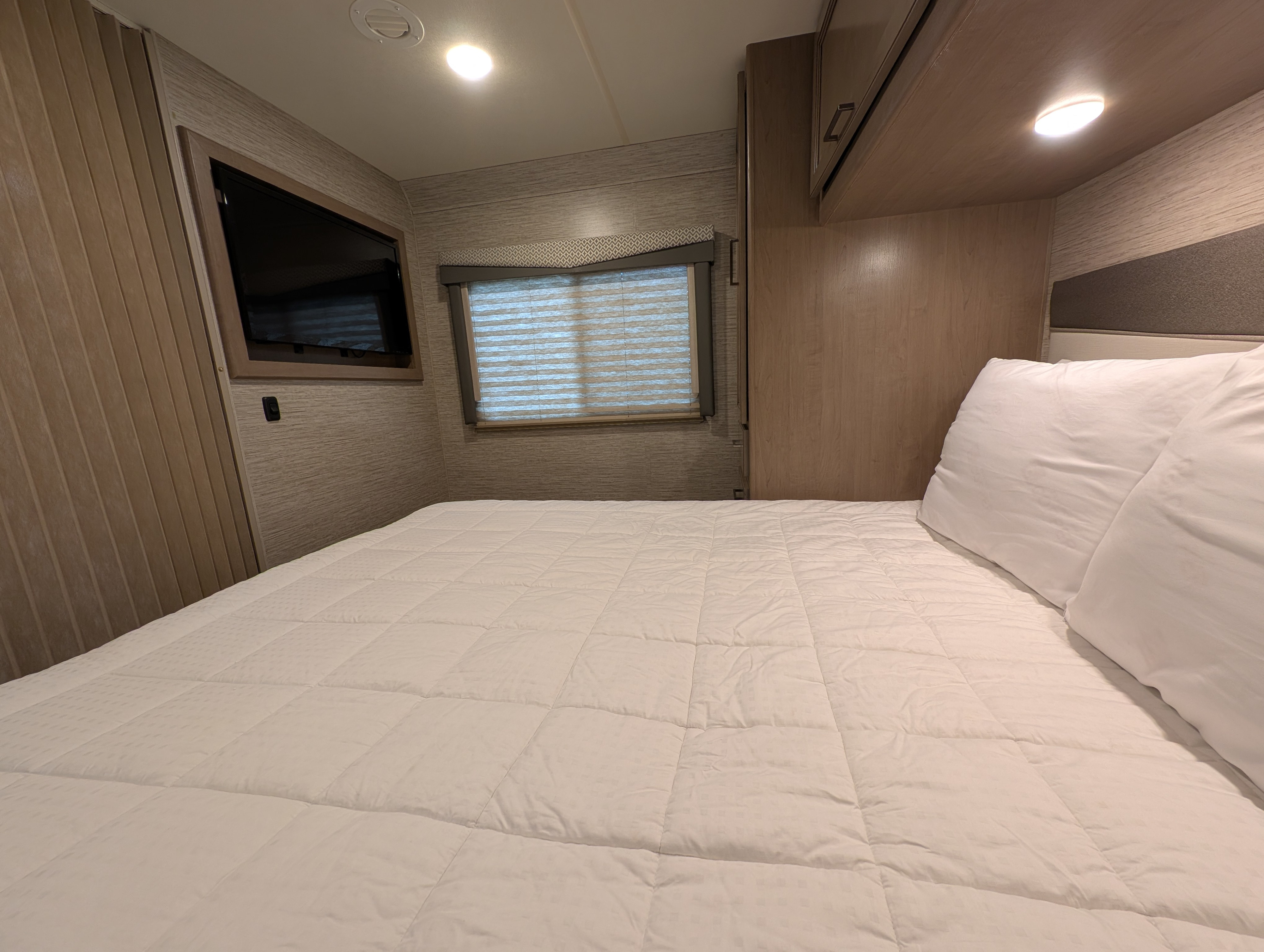 Cozy RV rental bedroom features a comfy bed, TV, window with blinds, and storage. Perfect for travel adventures.. Thor Four Winds 2021