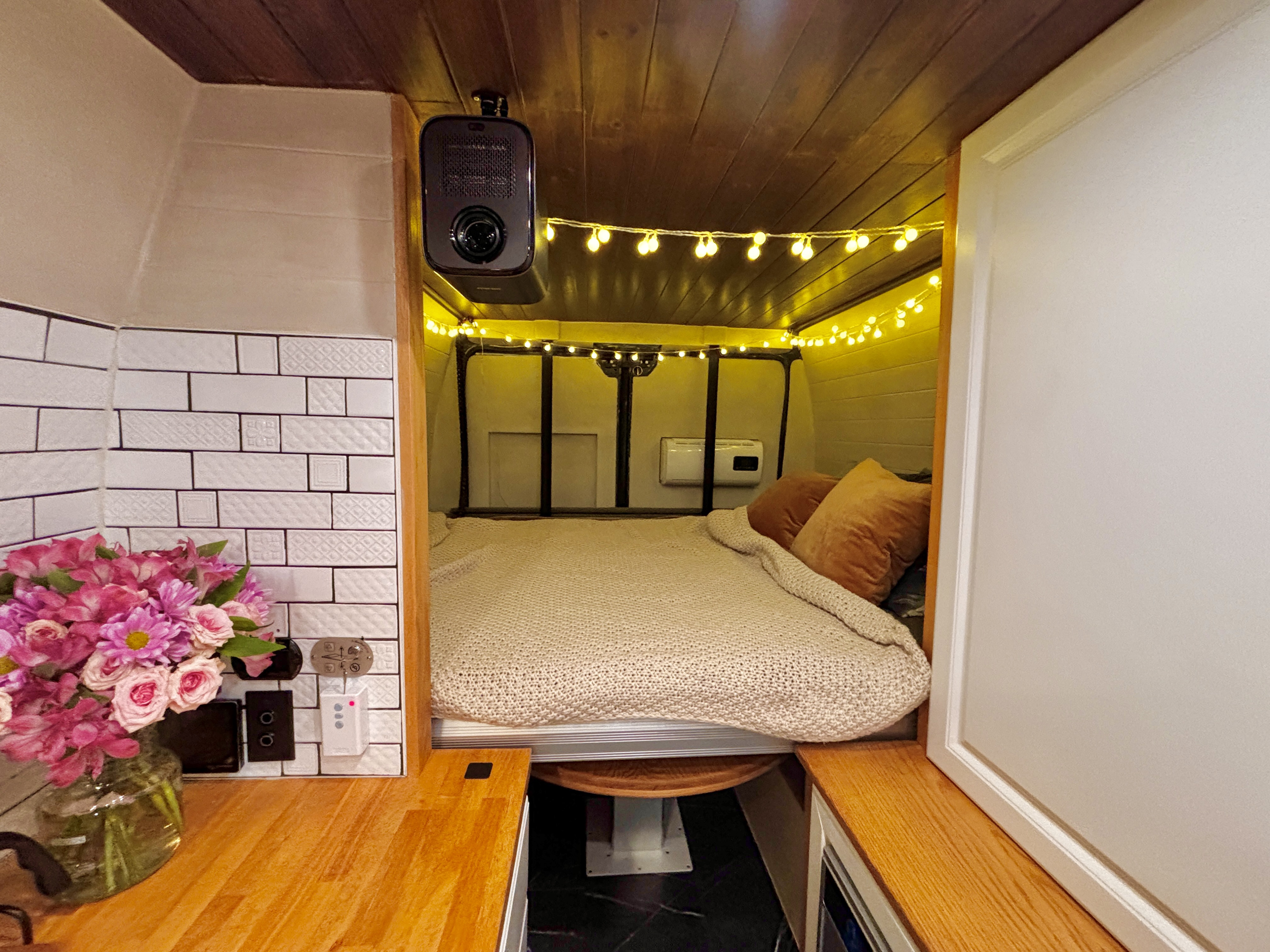 Step inside this cozy RV rental's bedroom, featuring bright string lights, a ceiling projector, and a compact kitchen.. Ram Promaster 3500 2019