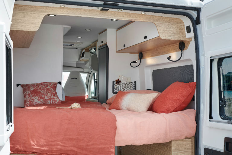 Discover this cozy RV rental's bedroom: coral bedding, wood accents, overhead storage, and reading lights.. Pilote V600G 2025