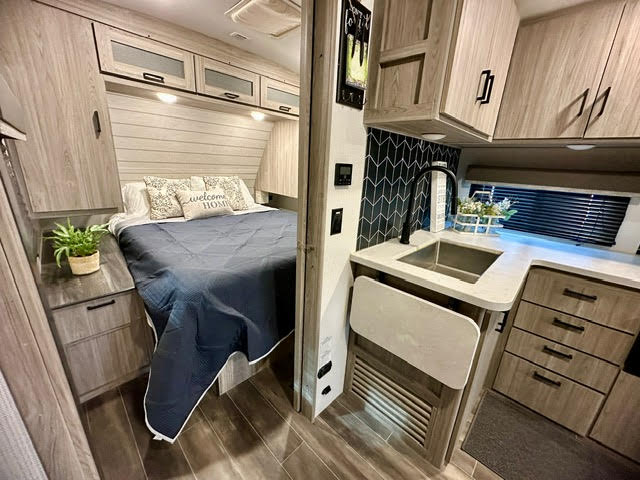 This RV rental interior showcases a cozy bedroom with light wood cabinetry and a modern kitchen with a sink and sleek tile backsplash.. Keystone Passport 2021