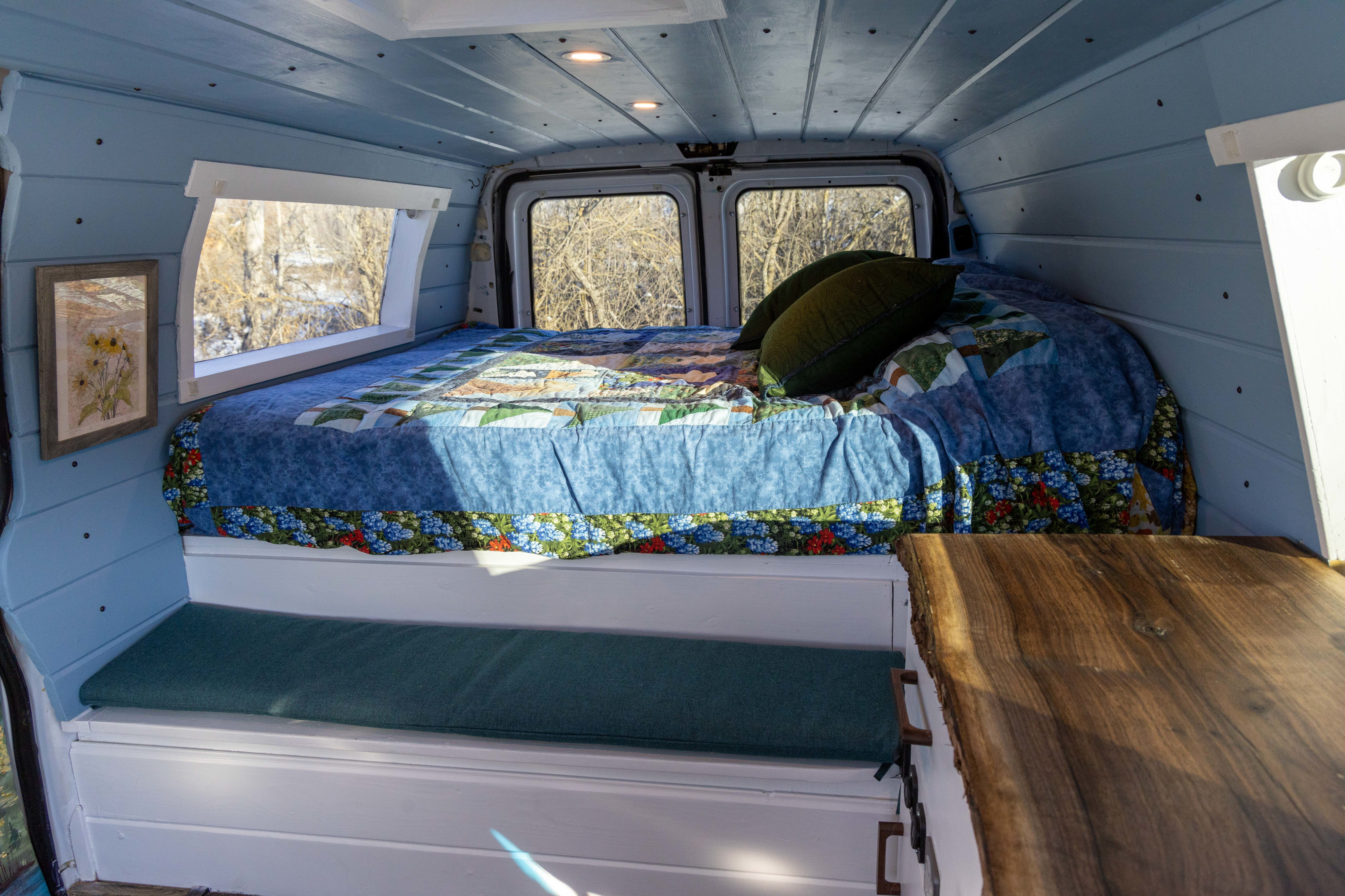 View inside a cozy RV rental: custom camper van with a bed, quilt, pillows, and built-in bench seating.. Ford Econoline 150 2009