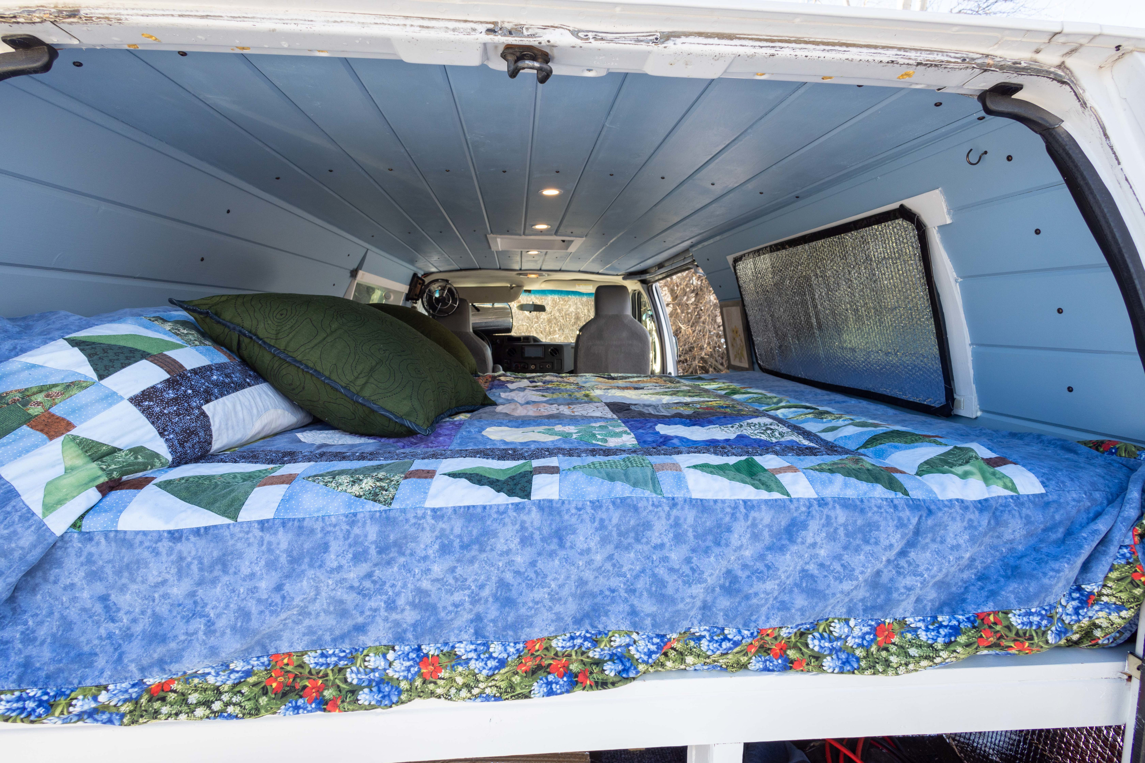 Inside an RV rental: a comfy bed with a quilted blanket & blue wood-paneled ceiling in a custom camper van.. Ford Econoline 150 2009