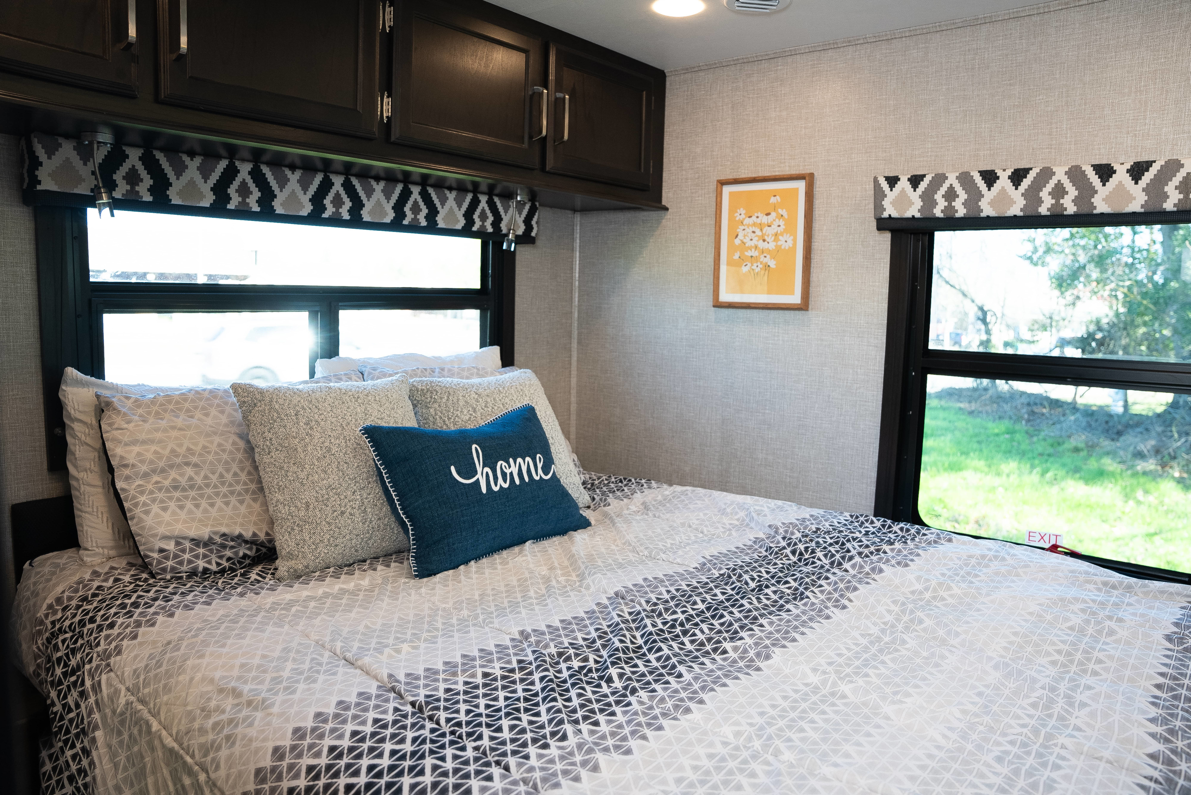 RV rental bedroom with a cozy bed, patterned bedding, "home" pillow, and large windows revealing outdoor greenery.. Entegra Coach Odyssey 2022