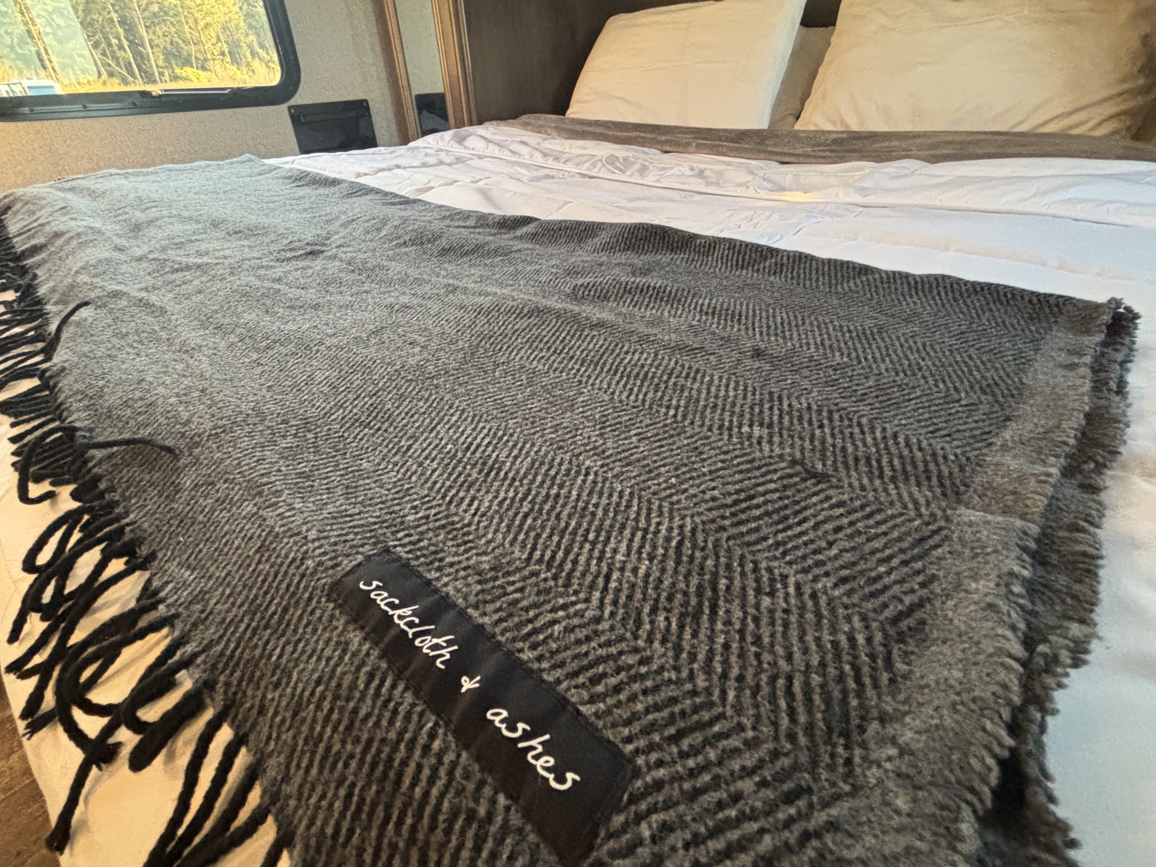 Cozy RV rental bed with a gray herringbone blanket, featuring fringe & a 'Sackcloth & Ashes' tag.. Keystone RV Passport Ultra Lite 2019