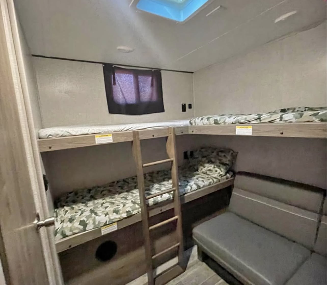 Inside an RV rental: bunk beds with camo bedding, a small ladder, window, and a gray couch. Compact sleeping and lounging space.. Crossroads RV Zinger 2021