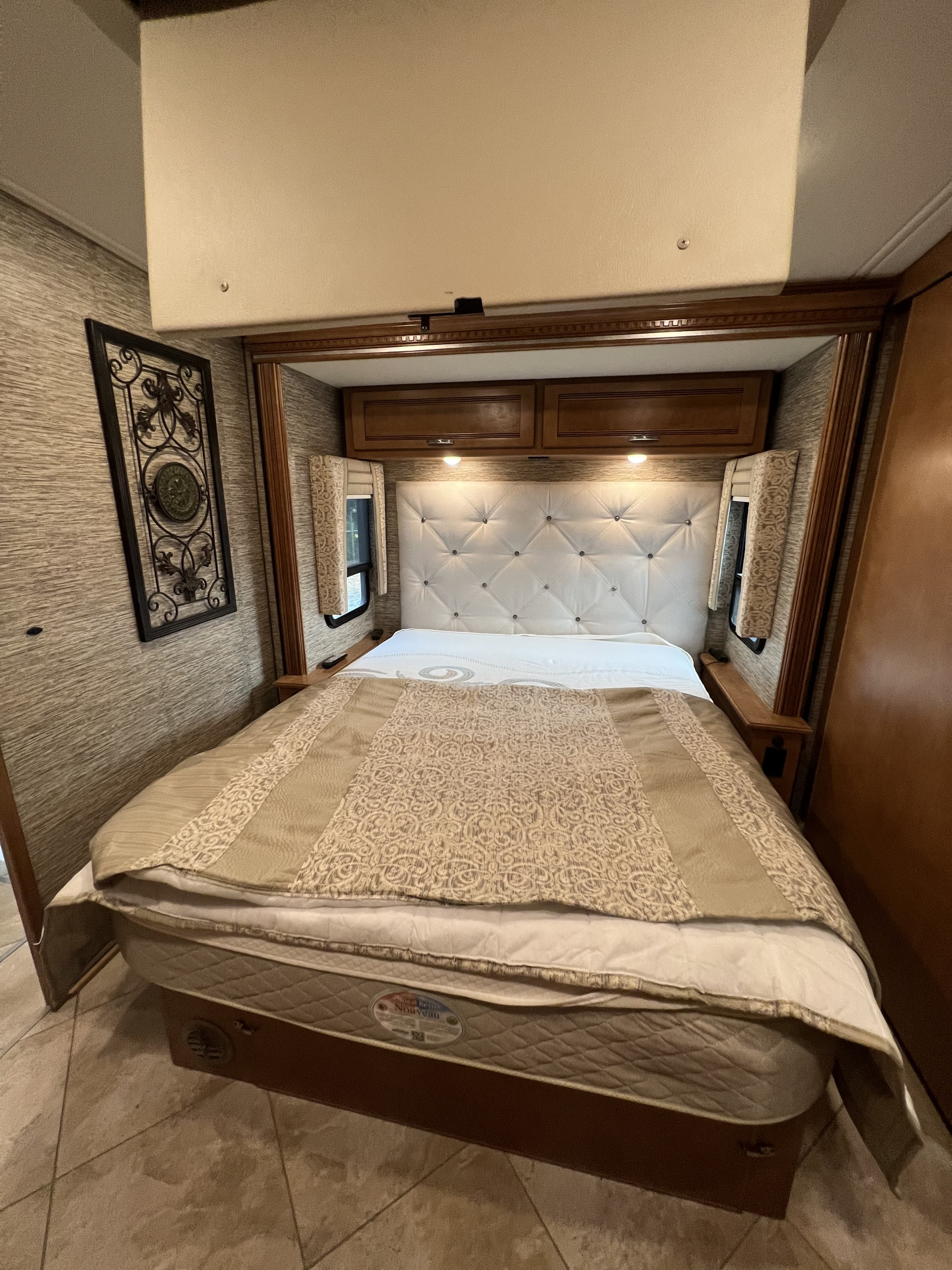 Comfortable RV rental bedroom featuring a large bed with a tufted headboard, cabinetry, and decorative accents.. Winnebago Forza 2019