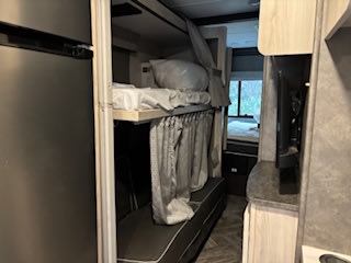 Inside an RV rental: a dark fridge, central bunk beds with curtains, and a TV on a counter. Compact travel space.. Forest River Sunseeker LE 2022