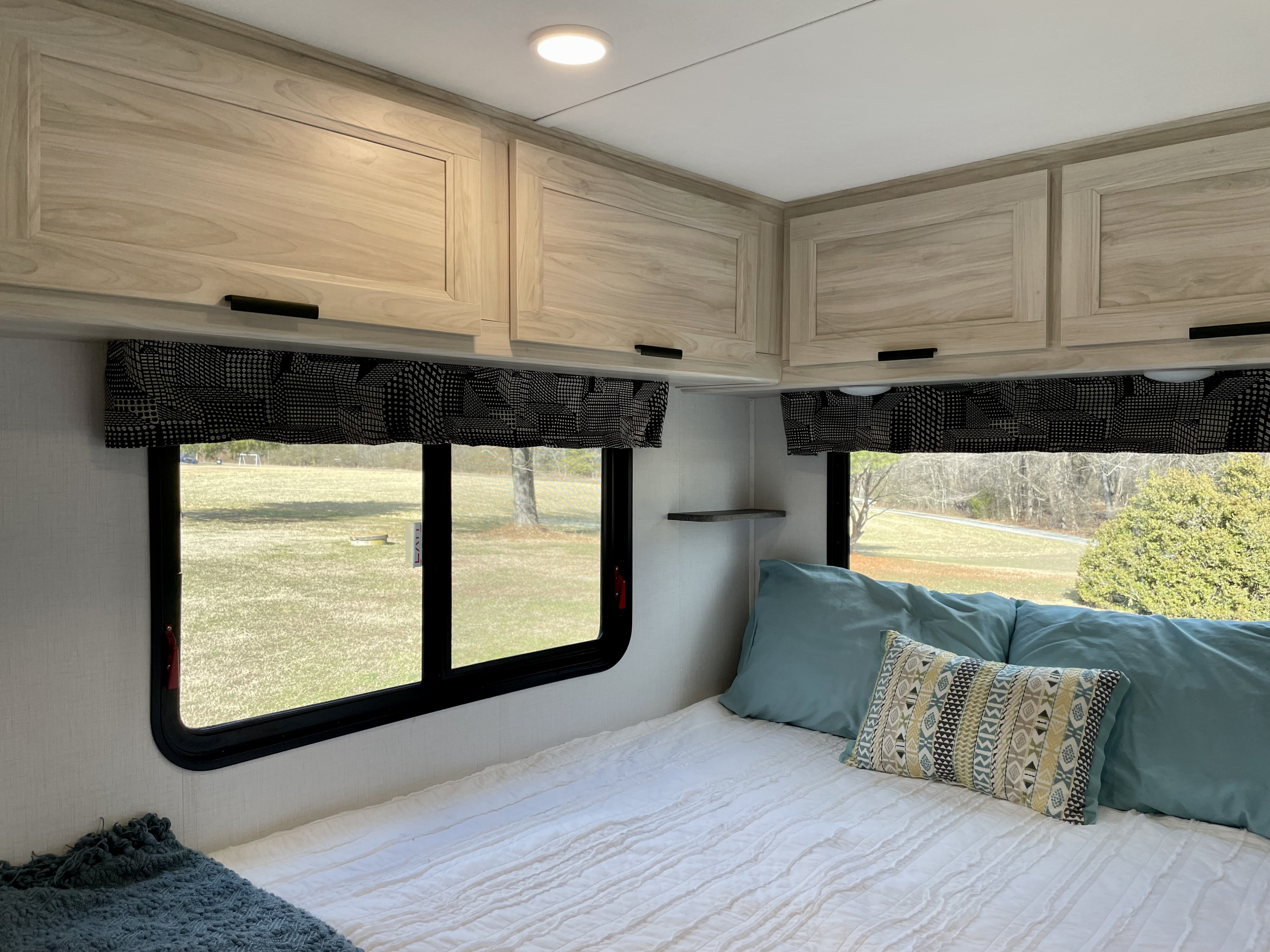 Bright RV rental bedroom features light wood overhead storage, windows with patterned valances, and a cozy bed.. Forest River Sunseeker LE 2023