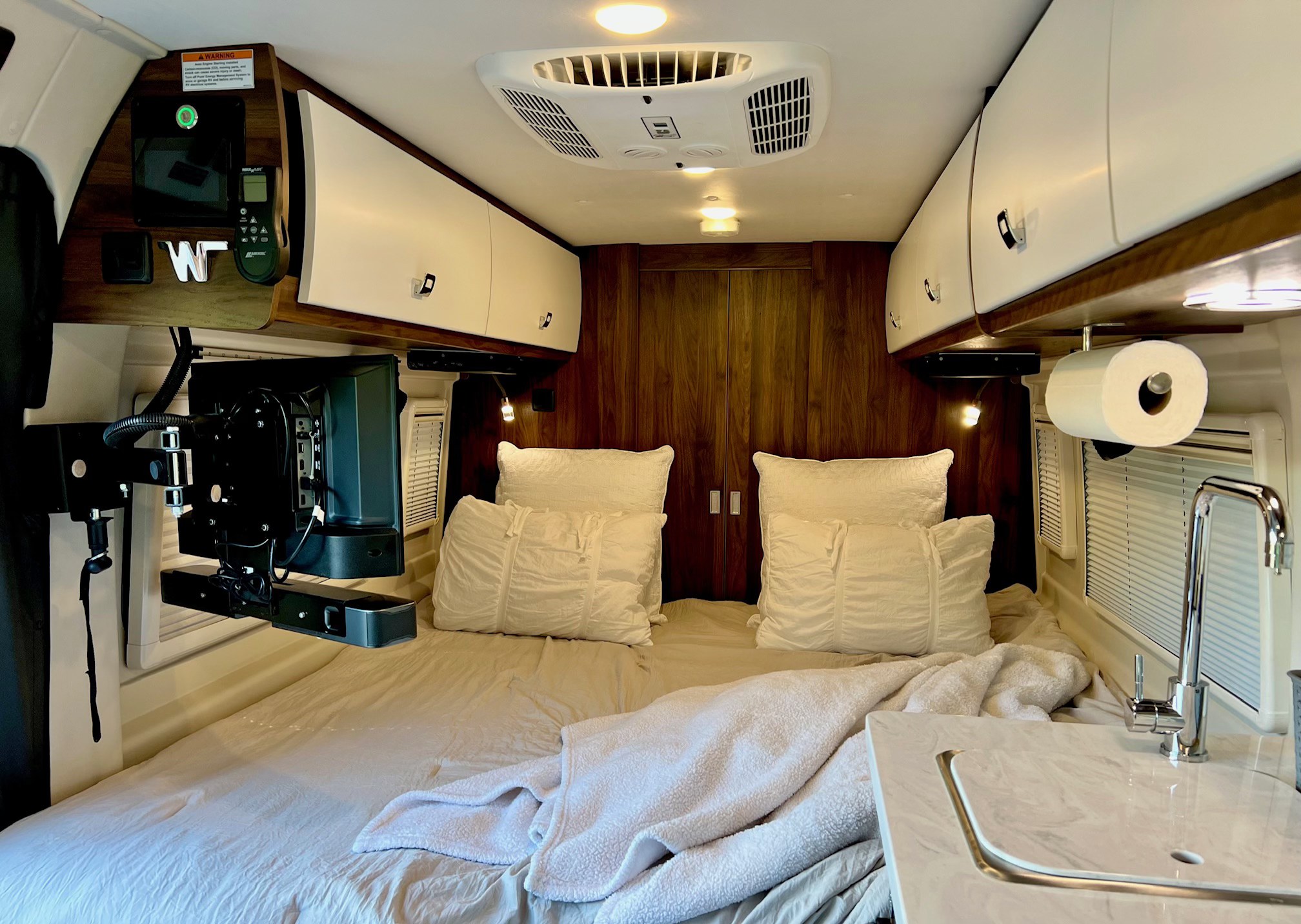 Inside this modern RV rental, a compact bedroom features a bed, TV, AC, overhead storage, and a small sink area.. Winnebago Travato 2023