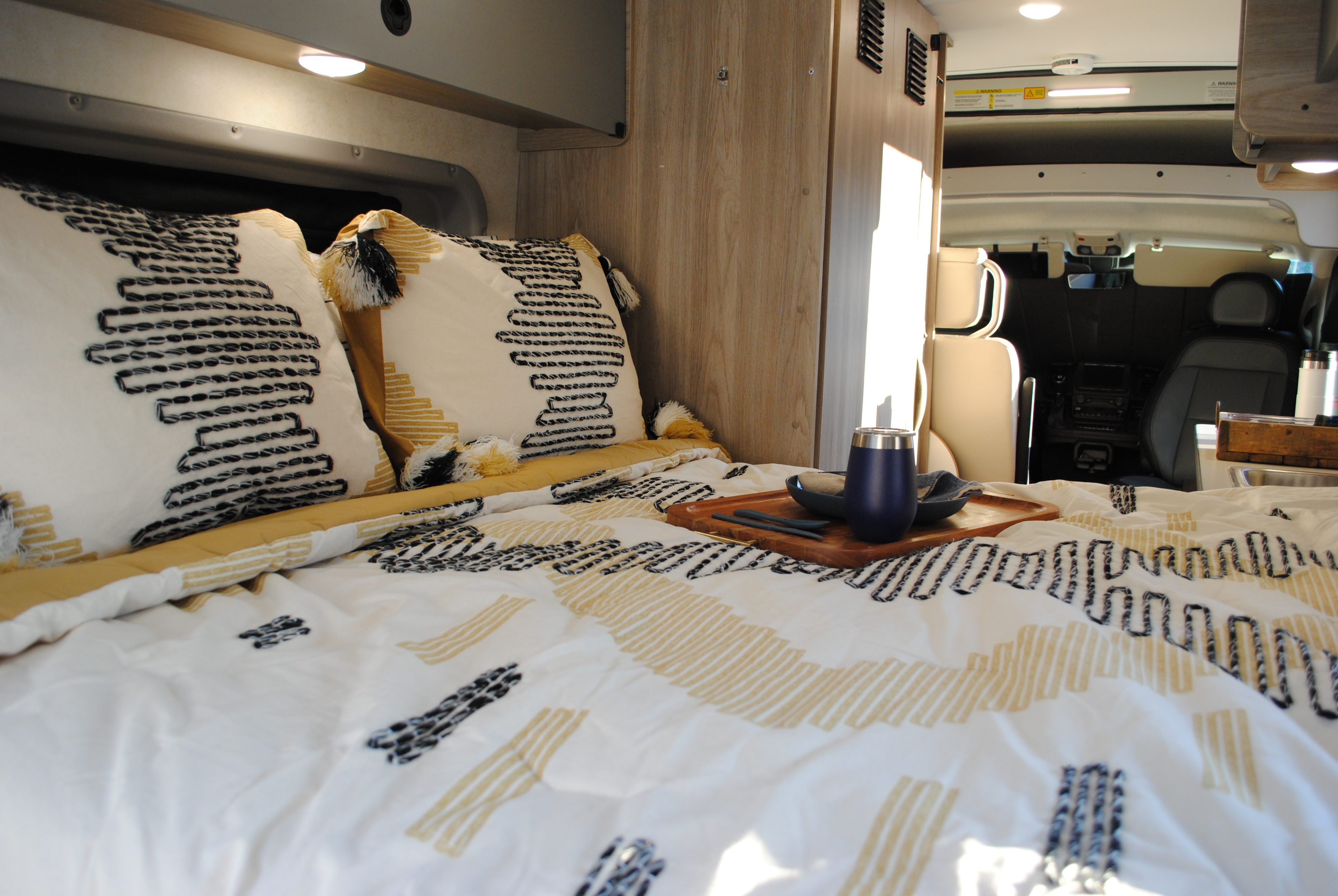 Inside a comfy RV rental: a bed with patterned bedding, decorative pillows, and a wooden serving tray. Front cab visible.. Winnebago Solis 2023
