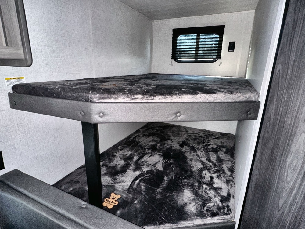 Inside an RV rental: compact bunk beds with dark grey mattresses, offering two cozy sleeping areas. A small window with blinds.. Keystone RV Hideout Sport 2024