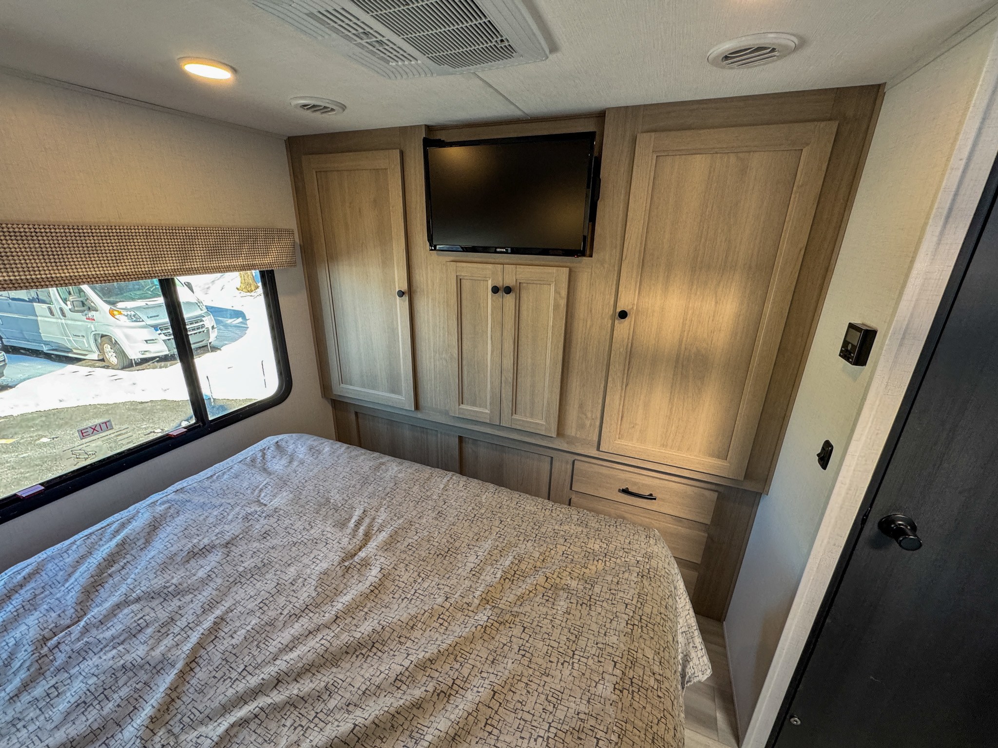 An RV rental bedroom features a patterned bed, light wood cabinetry, mounted TV, and a window looking out onto a snowy lot.. Forest River Sunseeker 2025