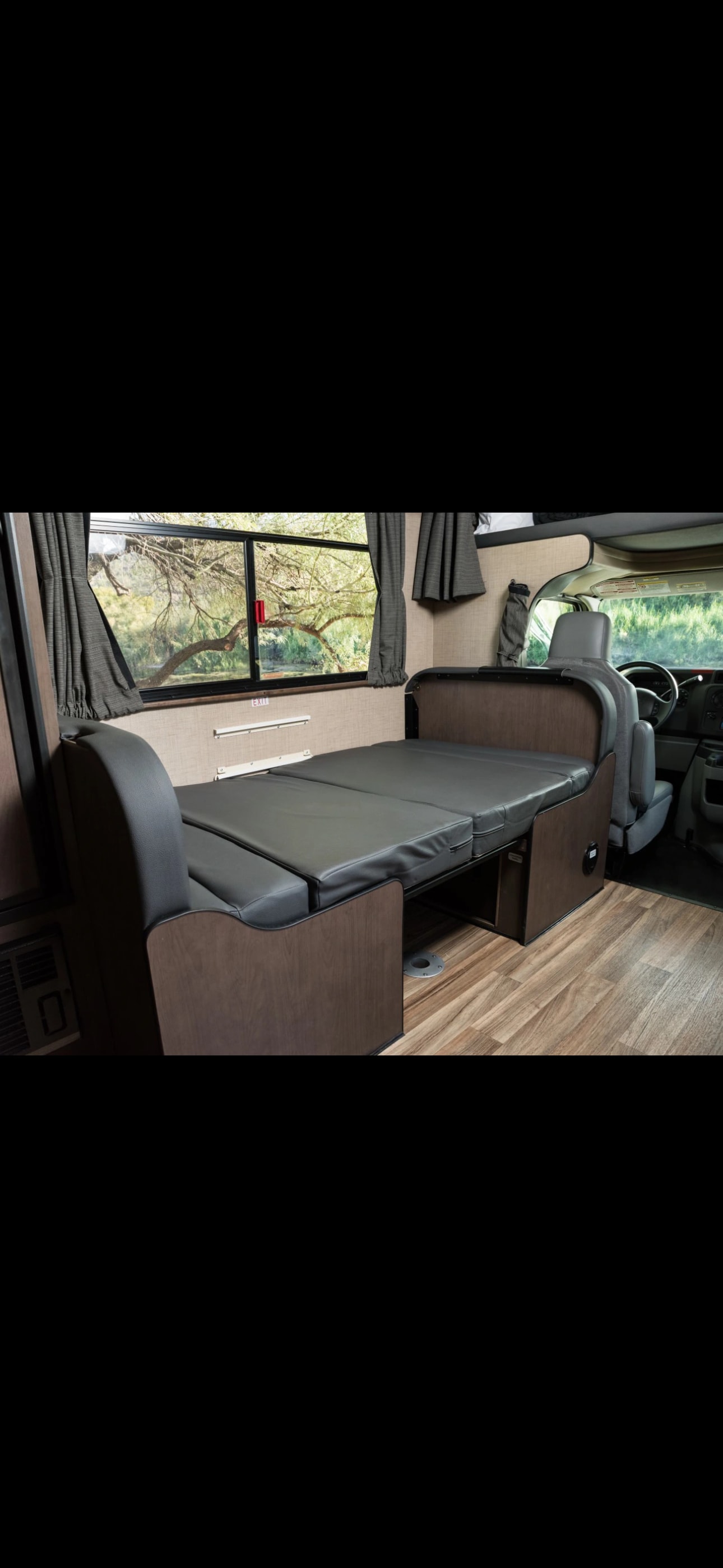 RV rental interior shows a flexible space: a dark grey cushioned bed/seating area, wood floors, and a scenic window view.. Thor Majestic 2021