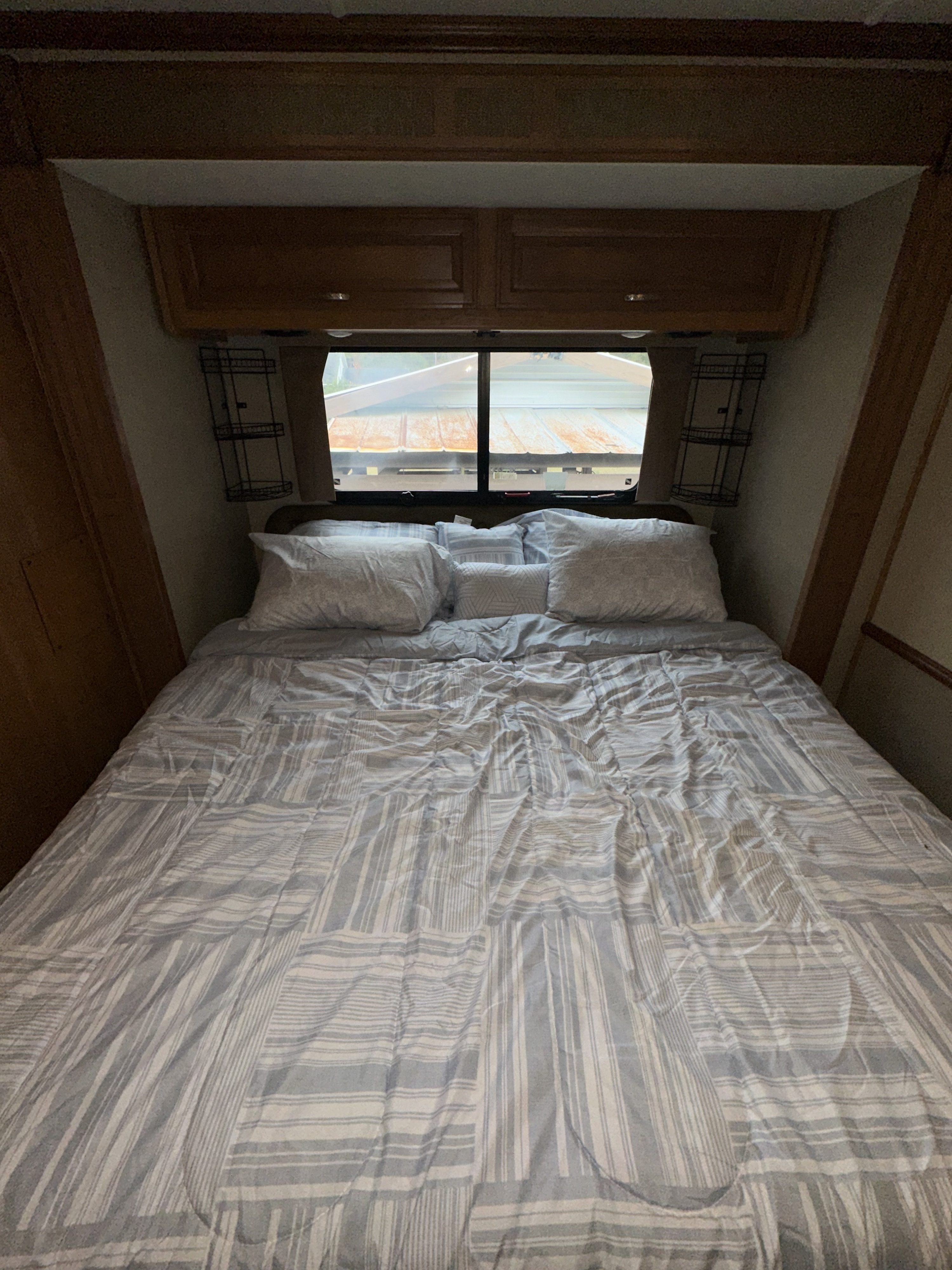 Cozy RV rental bedroom with striped bed, pillows, wooden cabinets, and window view. Perfect for travel.. Fleetwood Bounder 33C 2015