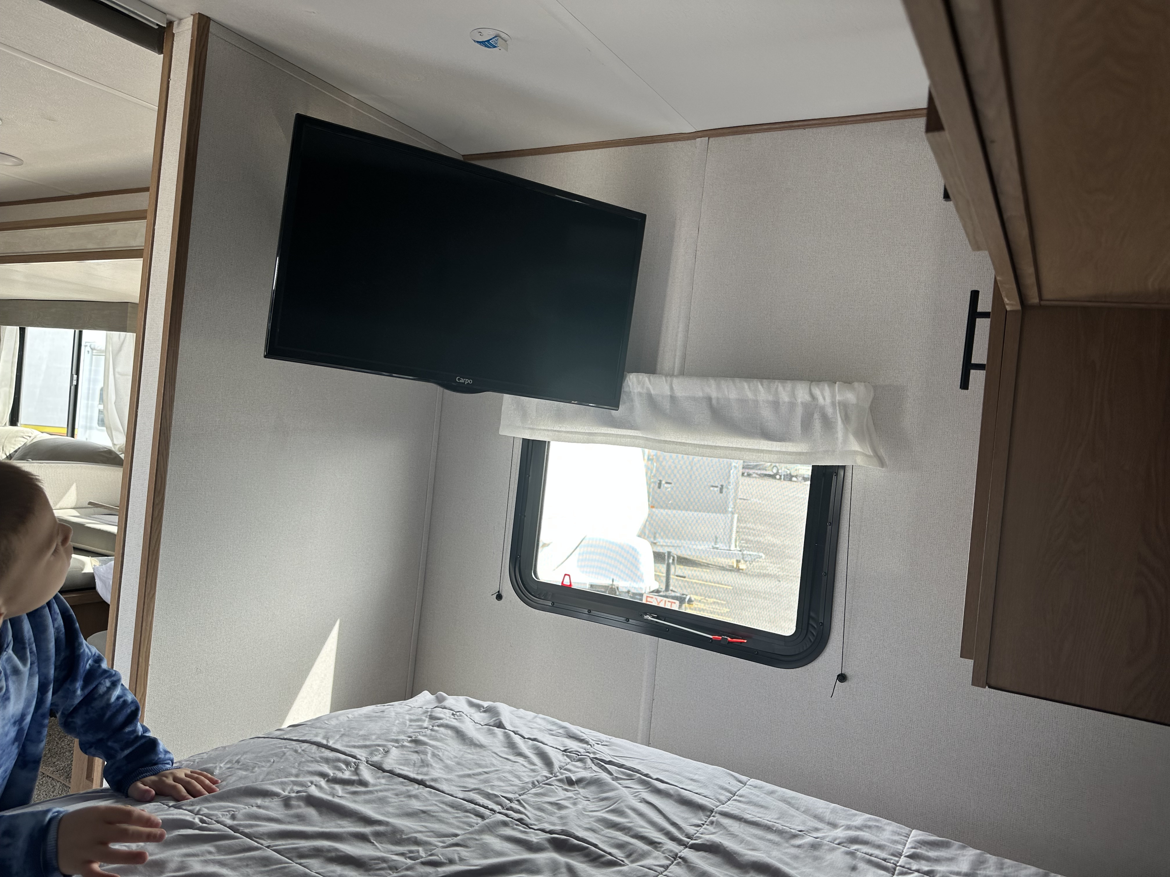 Inside a modern RV rental bedroom featuring a wall-mounted TV, comfy bed, and window with a child nearby.. Avenger Avenger Trailer 2022