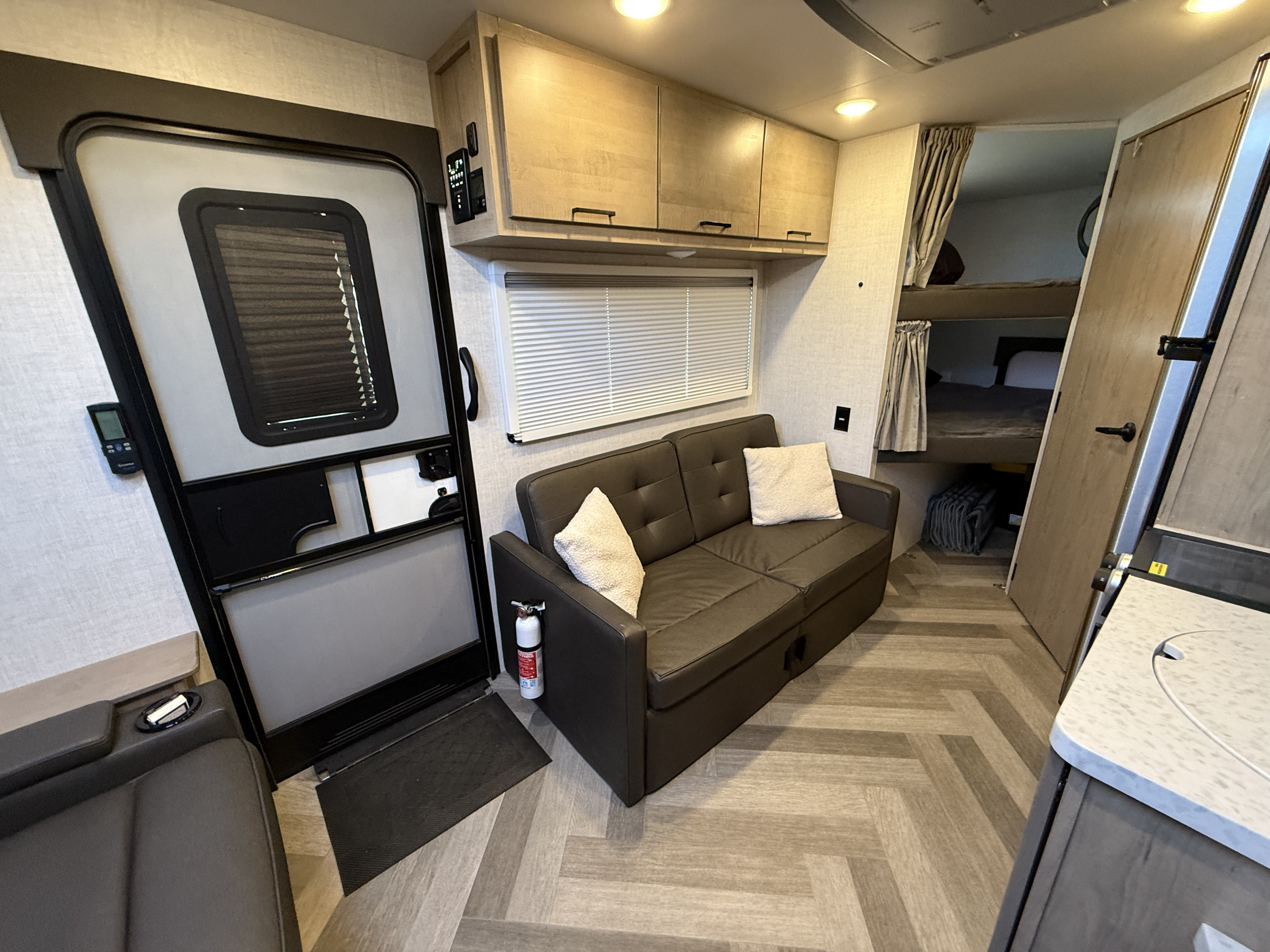 Inside an RV rental: view of the entry door, sofa, overhead cabinets, bunk beds, and a kitchen counter.. Ember Overland 2023